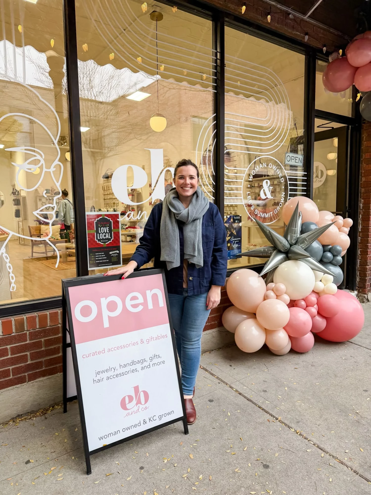 Woman standing outside a store with balloon decorations and an open sign, smiling, in front of large glass windows.