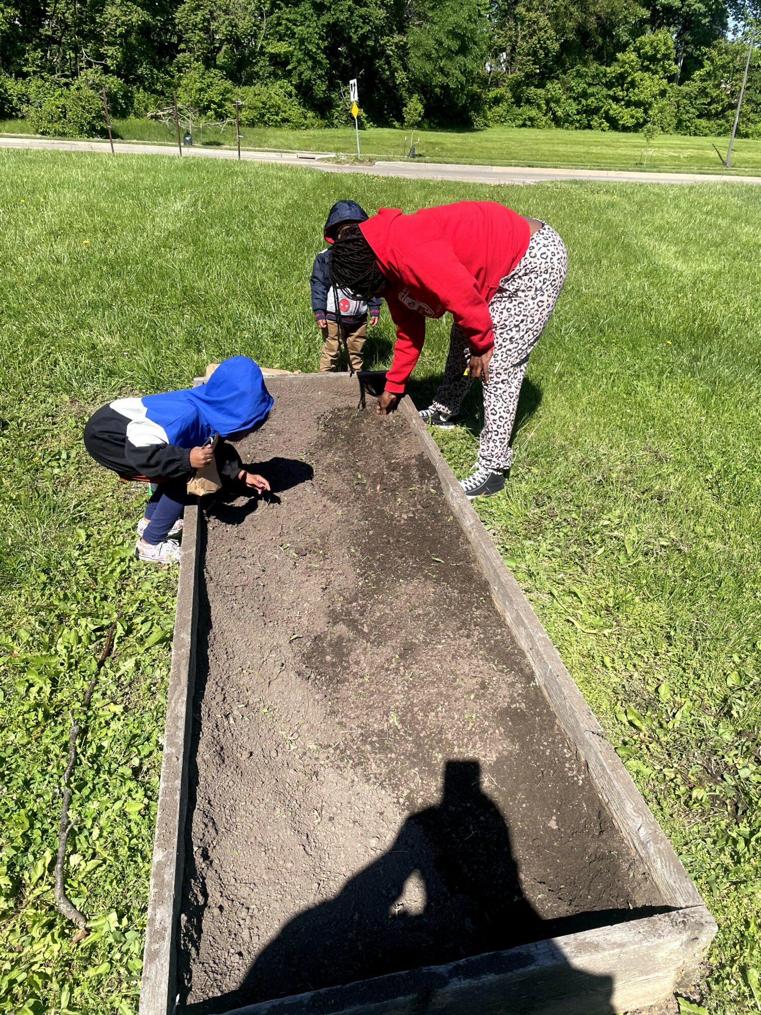 Two people, one child and one adult, planting or preparing soil in a rectangular garden bed outdoors on grass, with trees in background.