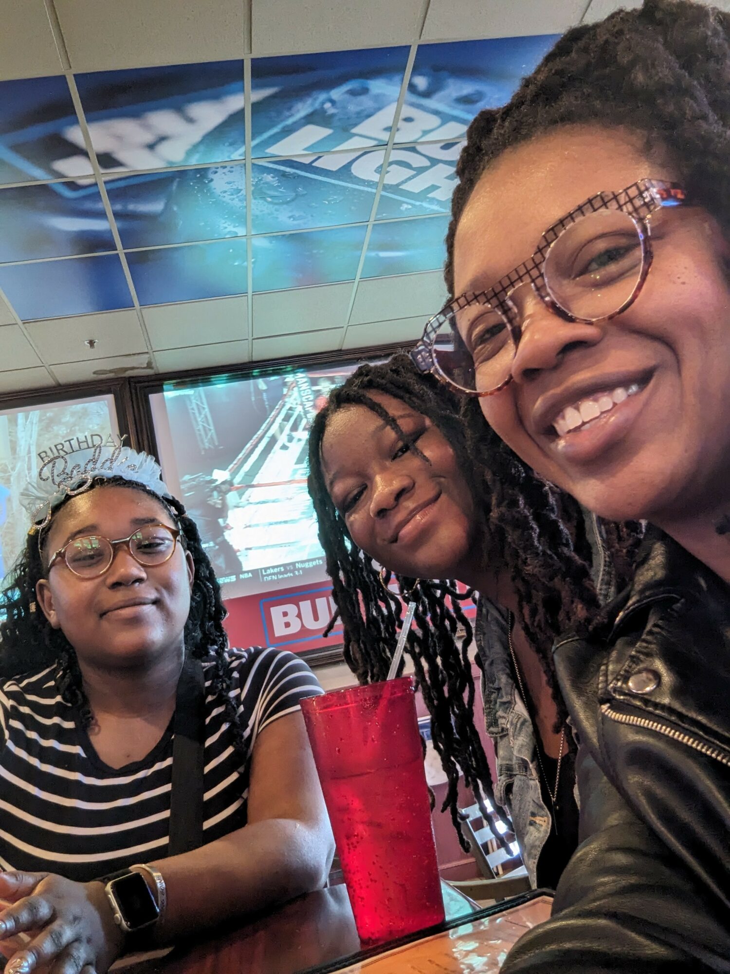 Three women smiling at a table with a red cup, in a fast-food restaurant with a digital menu and ceiling tiles.