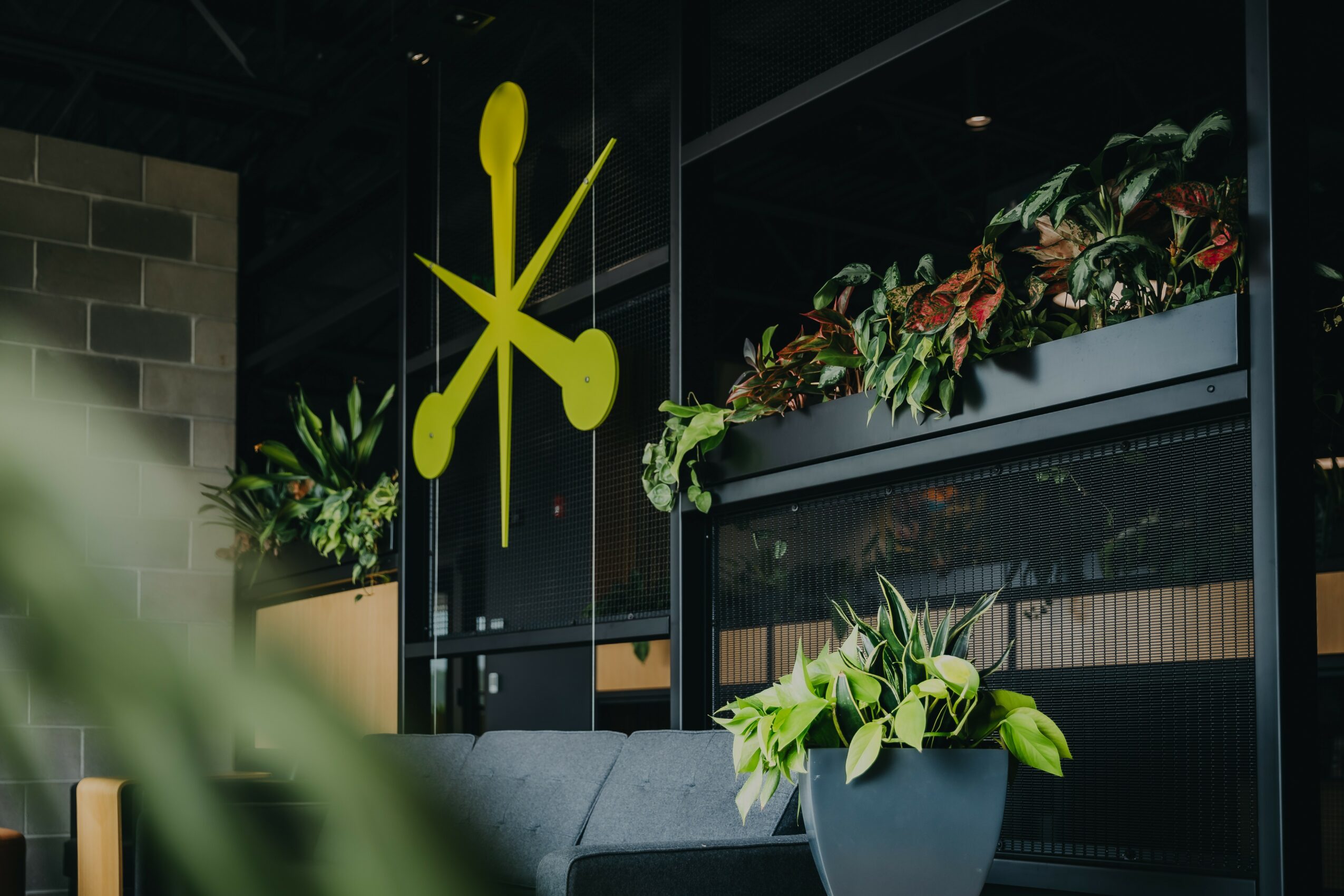 Indoor space with hanging yellow abstract art, potted plants, and a gray sofa, with a dark window and brick wall.