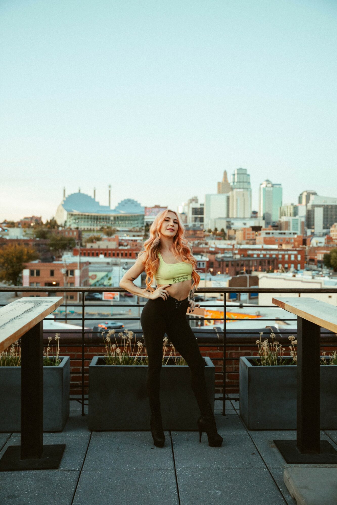 Woman with long hair standing on a rooftop balcony with city skyline in background, wearing a yellow top and black pants.