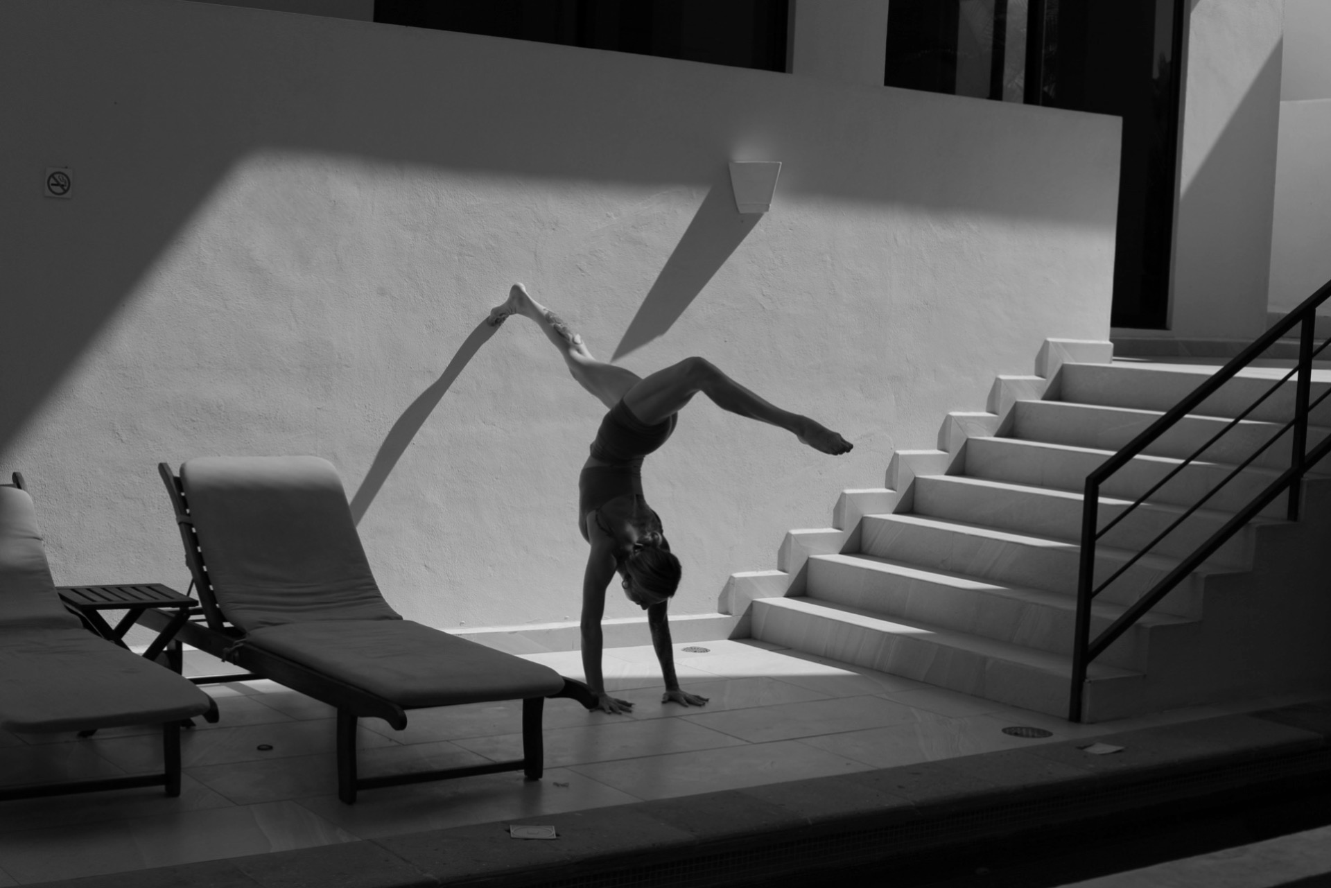 Person performing a handstand on a platform near stairs with a shadow cast on the wall, in black and white.