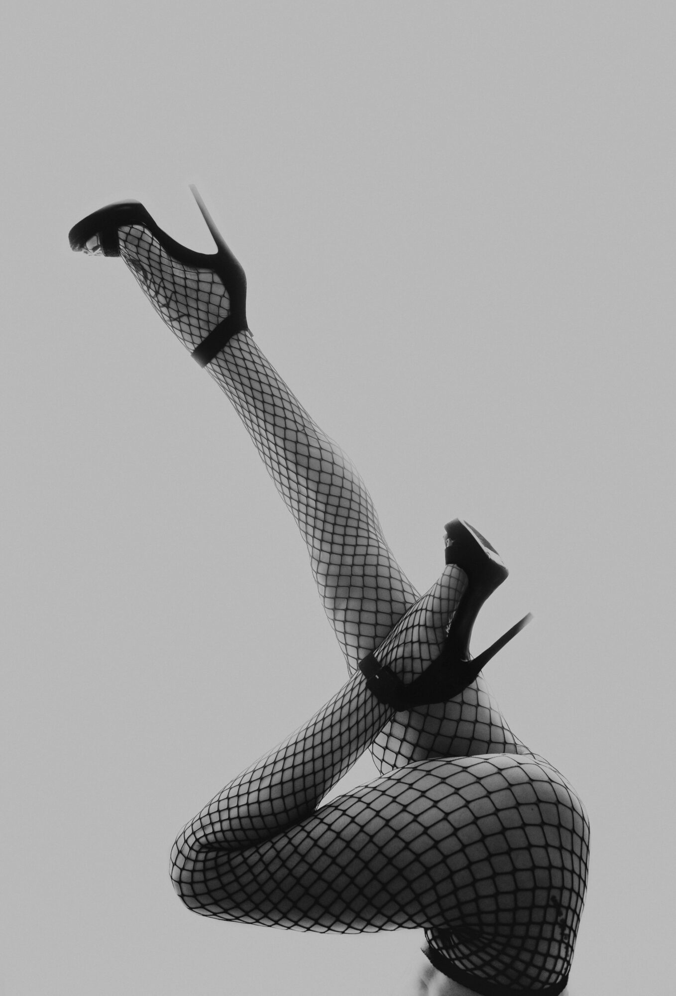 Person wearing fishnet stockings and high heels, with legs crossed and raised, against a plain background.