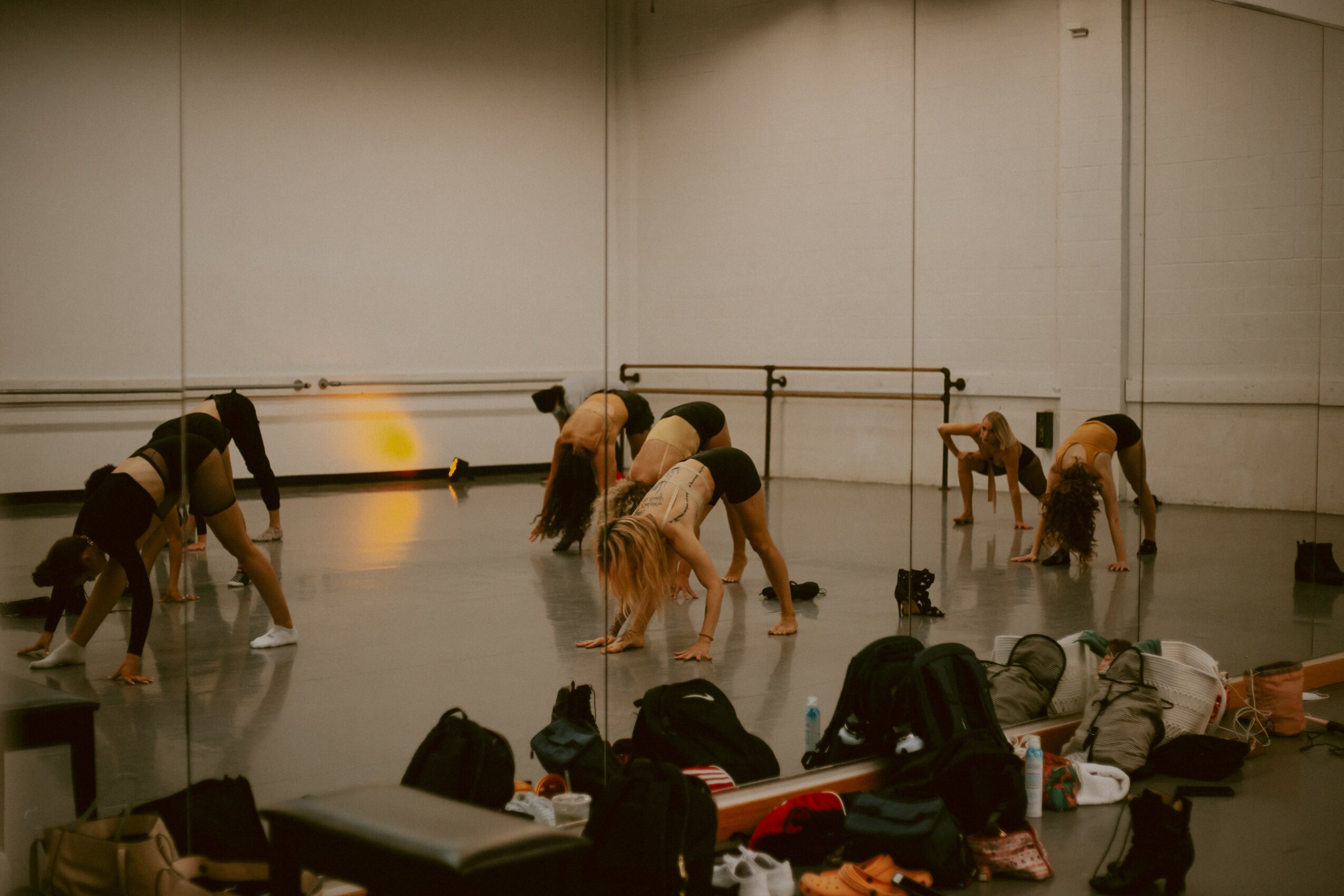 People in a dance studio bending forward, with bags and belongings on the floor in front, plain wall background.