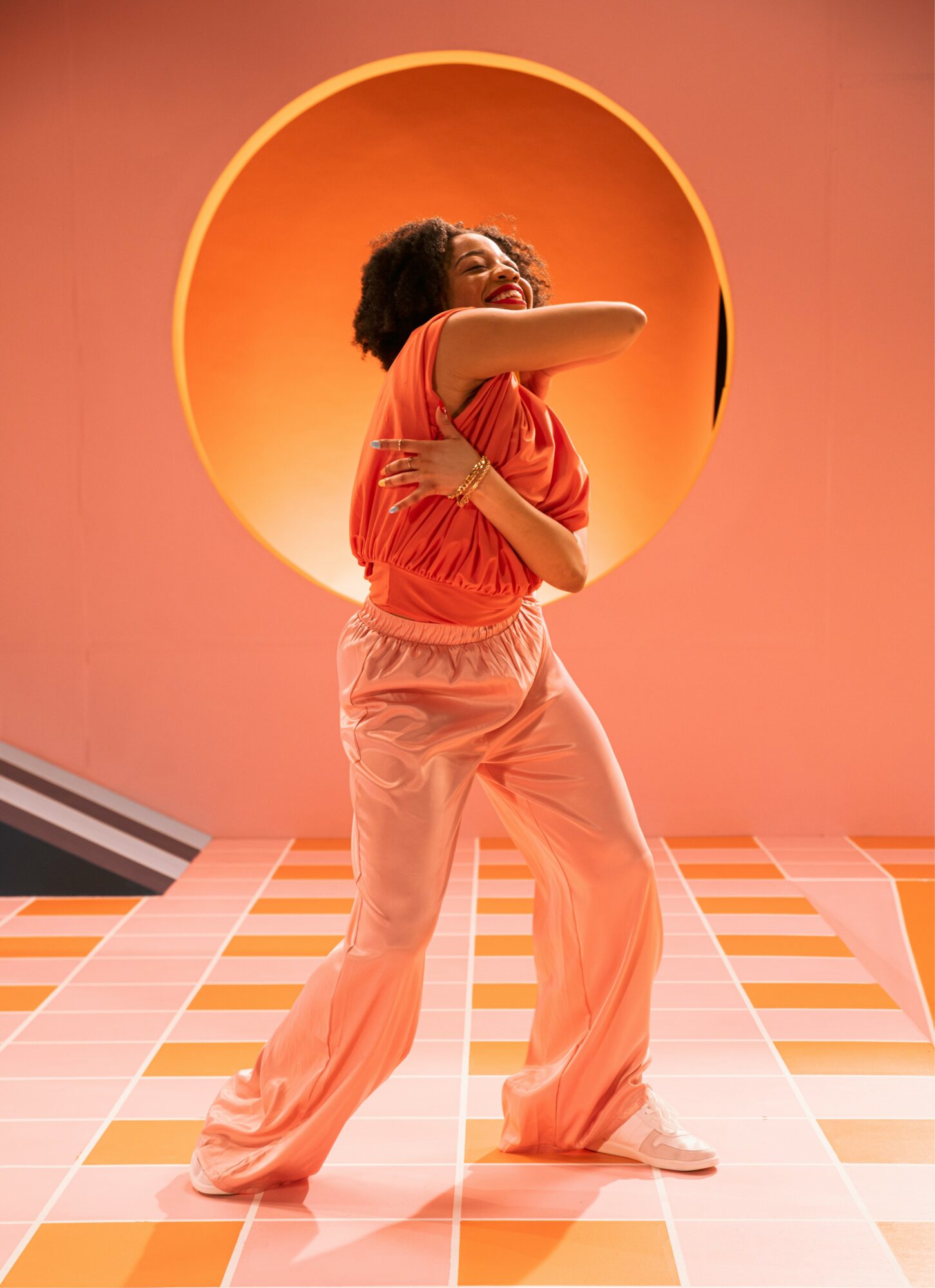 Person with curly hair in a red top and light-colored pants stands in front of a circular orange background, smiling and posing.