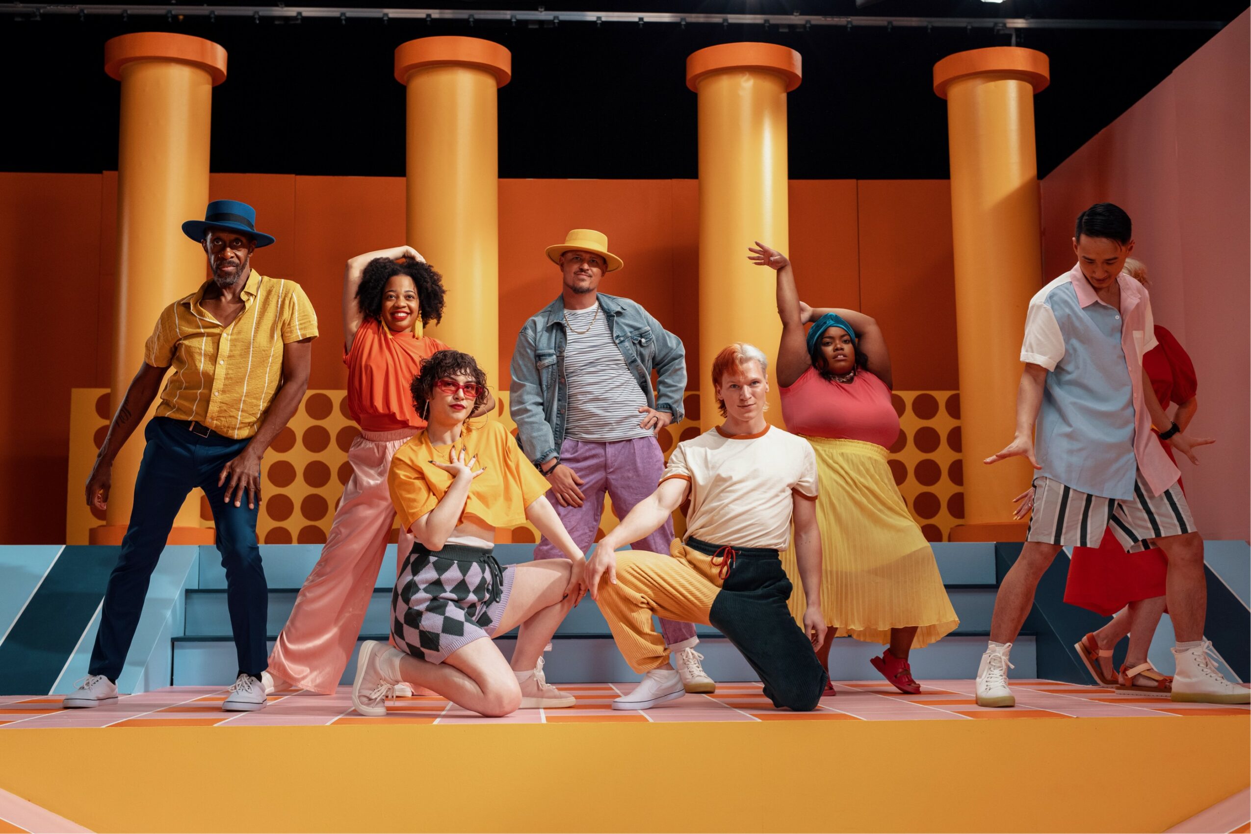 Group of seven diverse people dancing on colorful stage with orange columns in background.