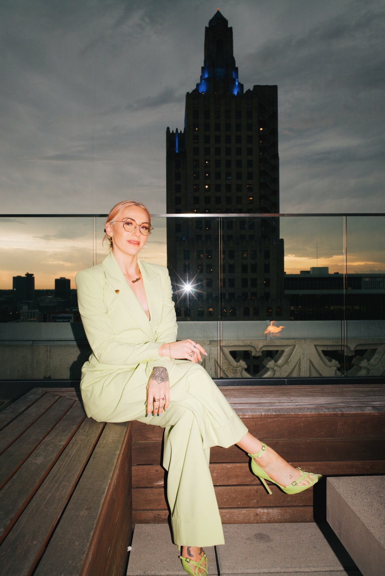 Woman in light-colored suit sitting on a bench on a rooftop with a tall building and cloudy sky in the background.