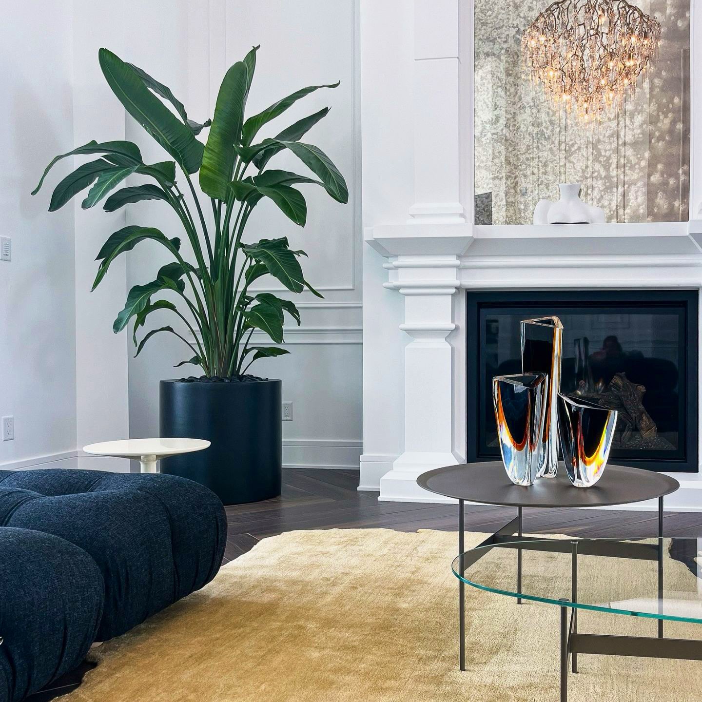 Living room with large potted plant, fireplace, glass vases on table, and a dark sofa, with a chandelier reflected in the window.