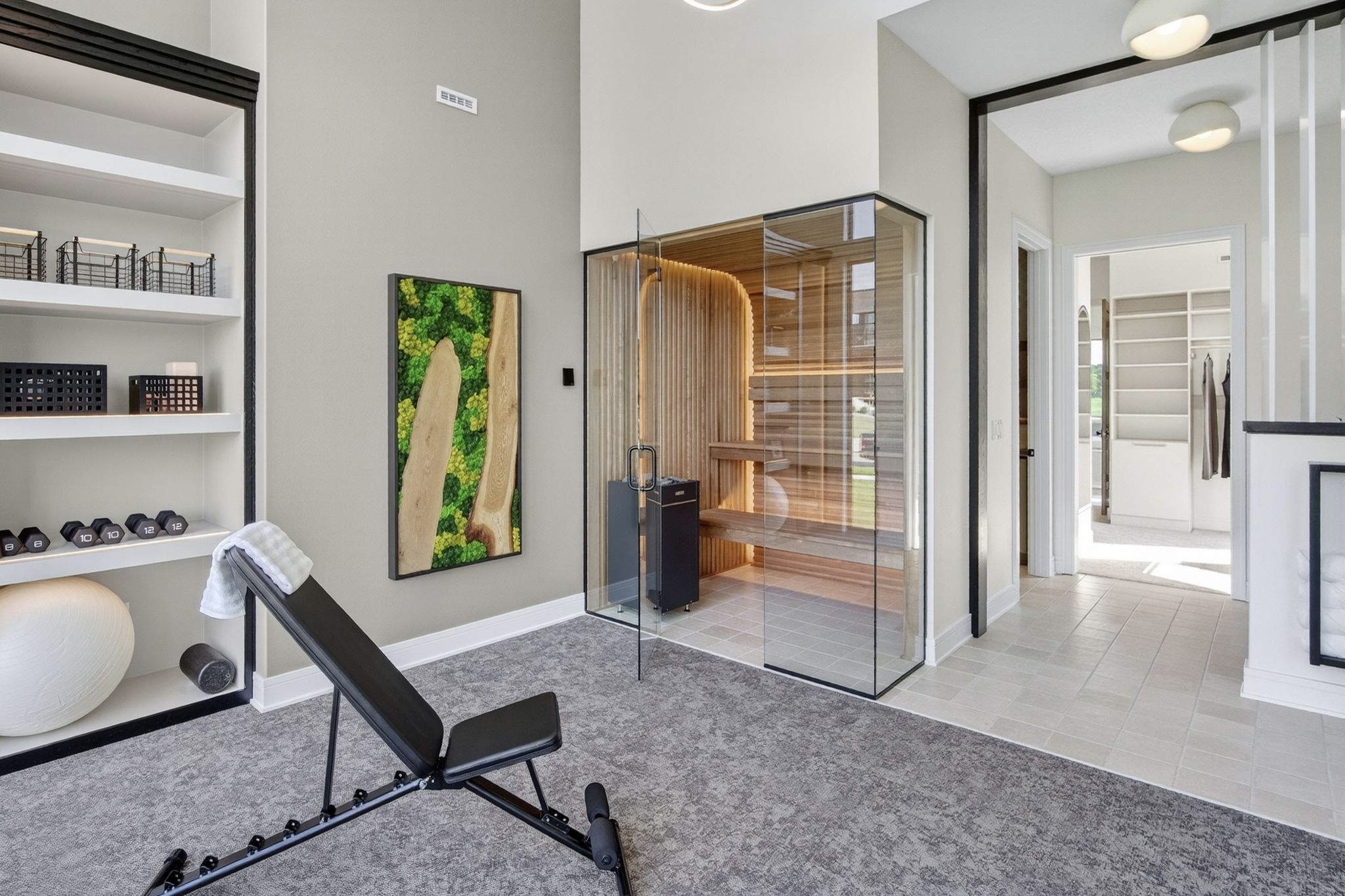 Modern interior with a glass sauna, bookshelf, artwork, and a black lounge chair on a gray carpet, bright natural light.