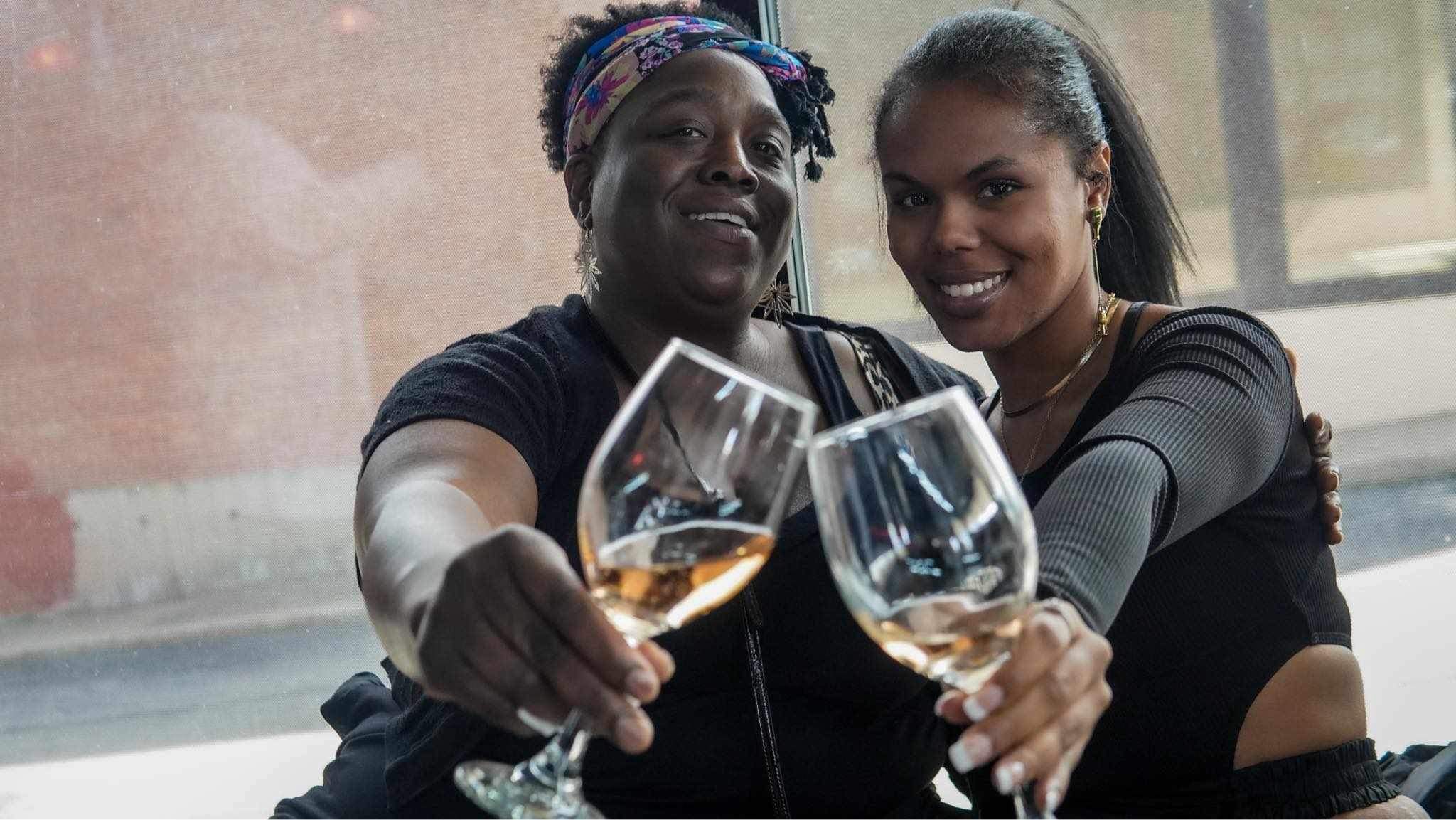 Two women clink wine glasses and smile at the camera indoors.