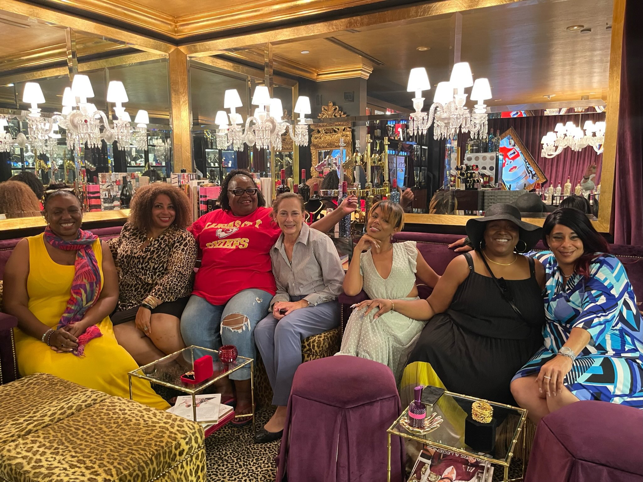 Seven women sitting on a sofa in a decorated room with chandeliers and mirrors, smiling at the camera.