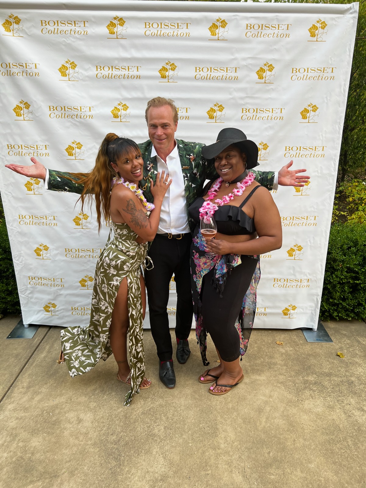 Three people standing outdoors in front of a white backdrop with gold text, smiling and posing with arms outstretched.