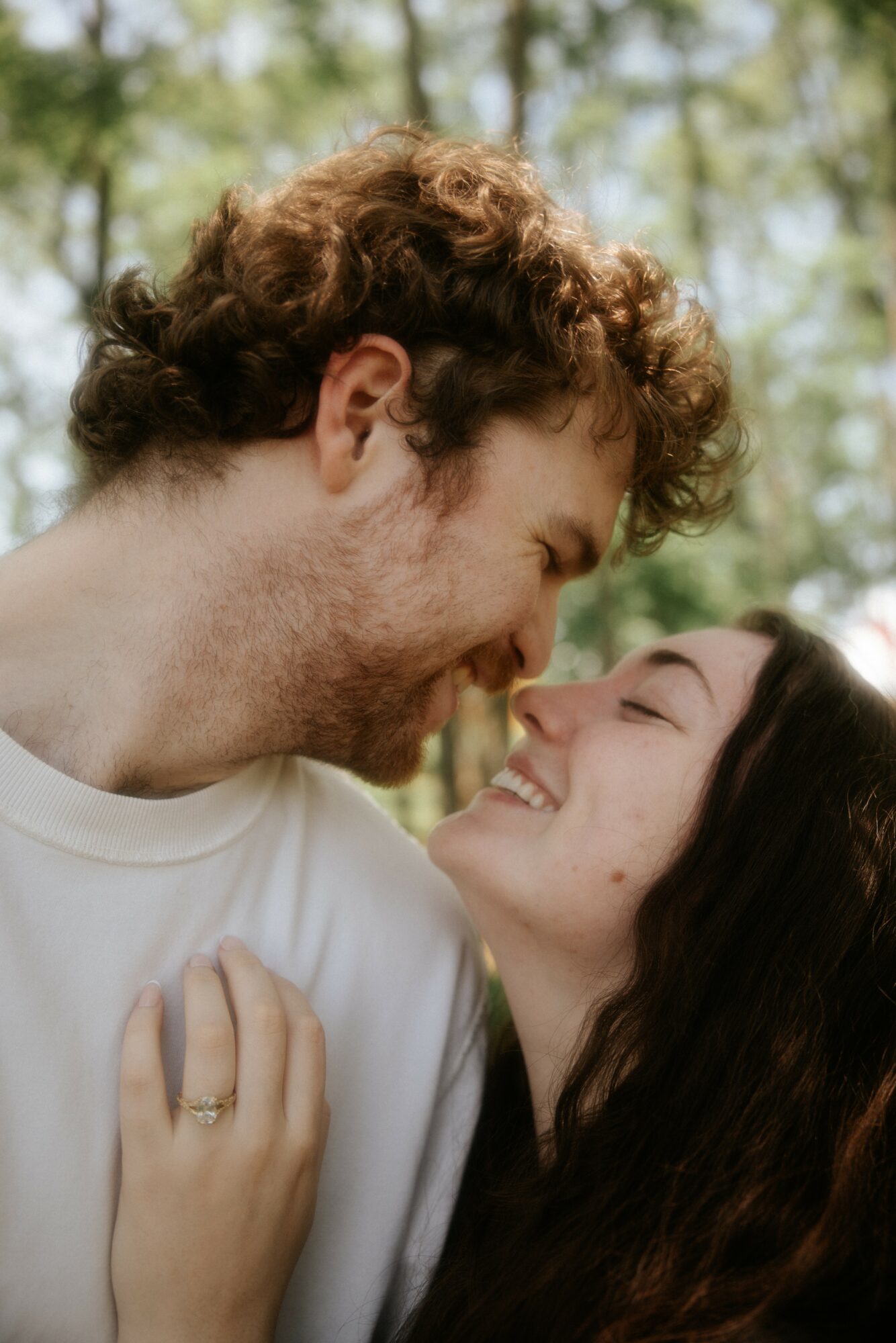 A man and woman smiling and looking at each other outdoors, close together, with trees in the background.