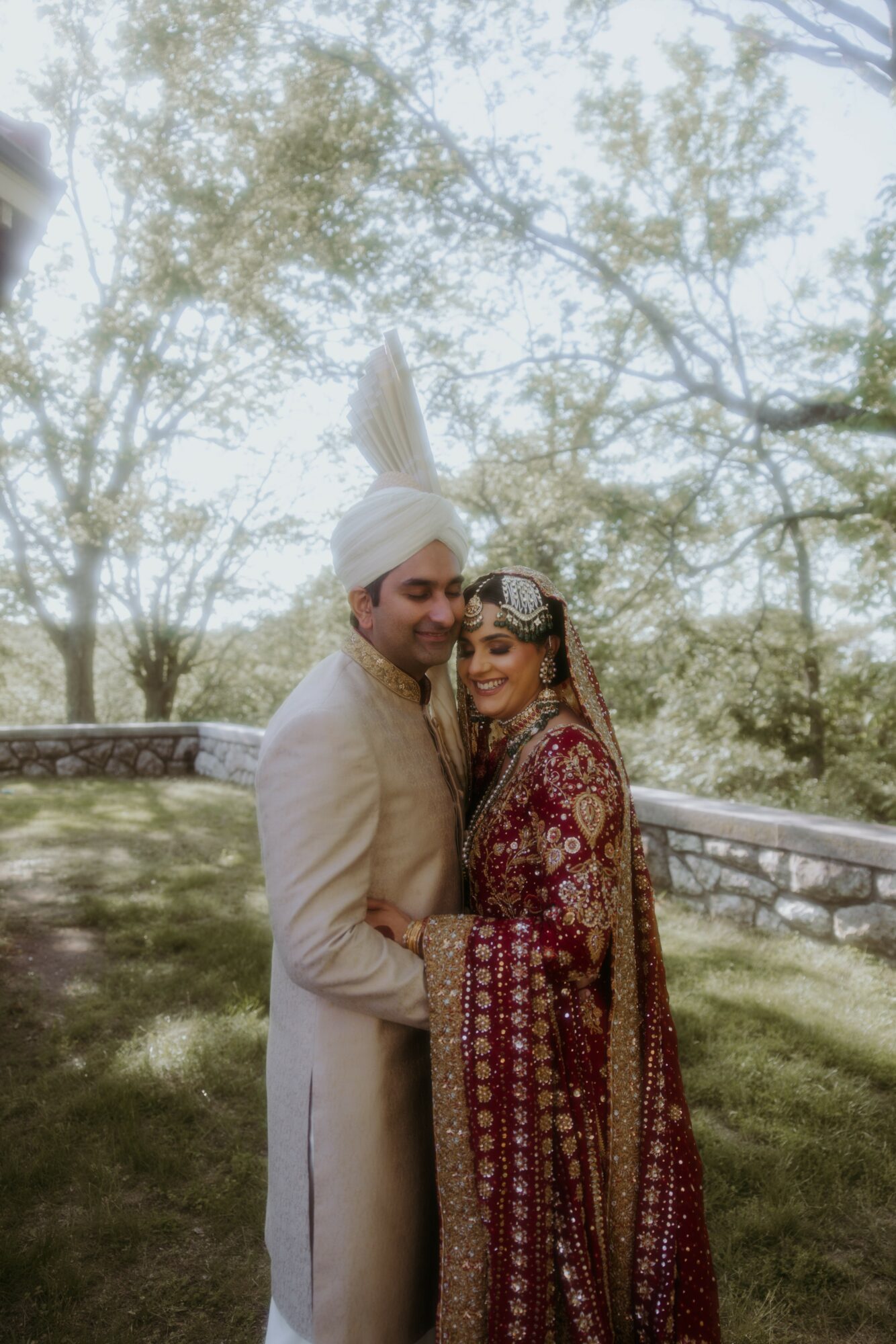 A couple in traditional wedding attire standing outdoors, smiling, with trees and a stone wall in the background.