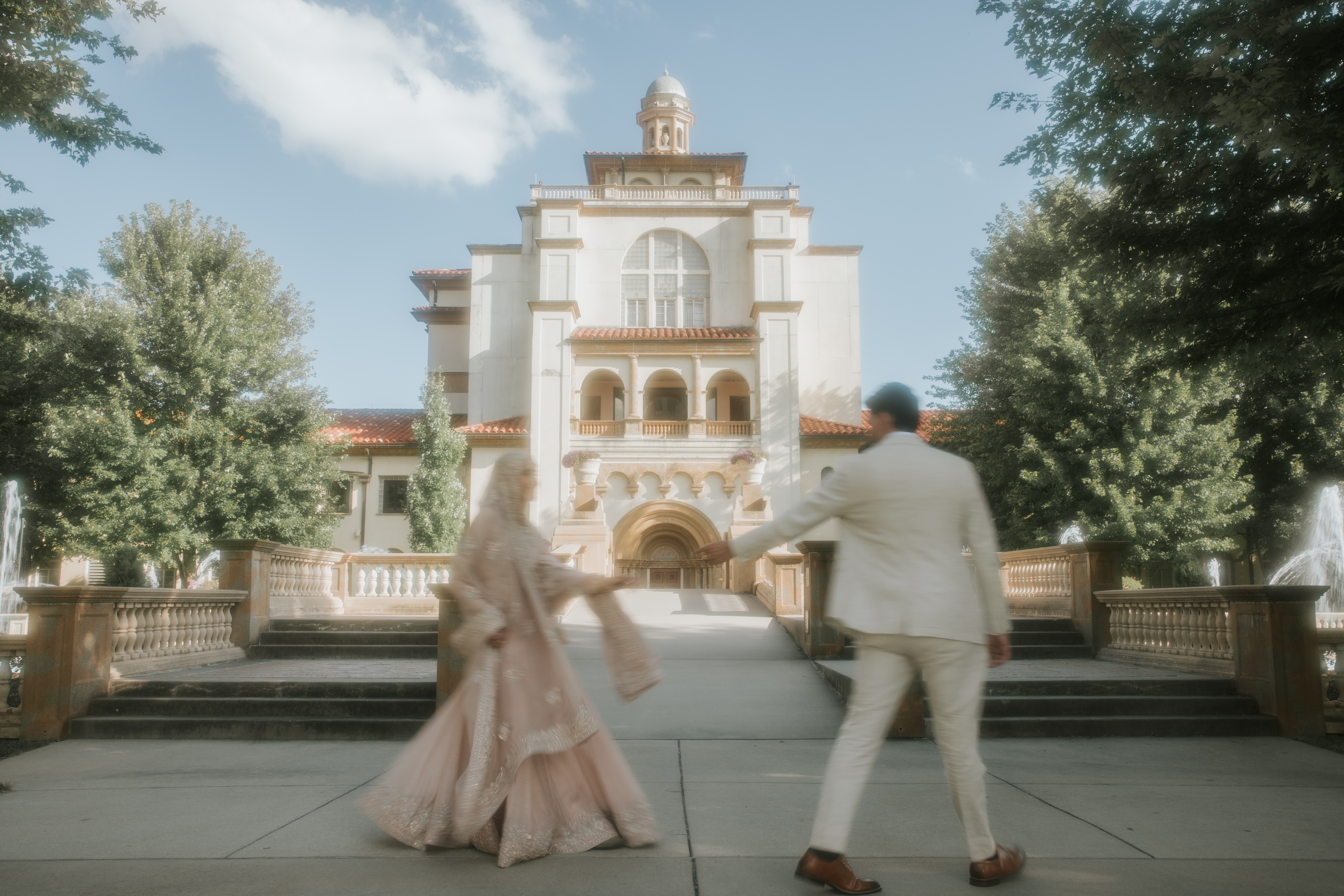 Two people in formal attire walk toward a large, ornate building with trees on both sides, under a partly cloudy sky.