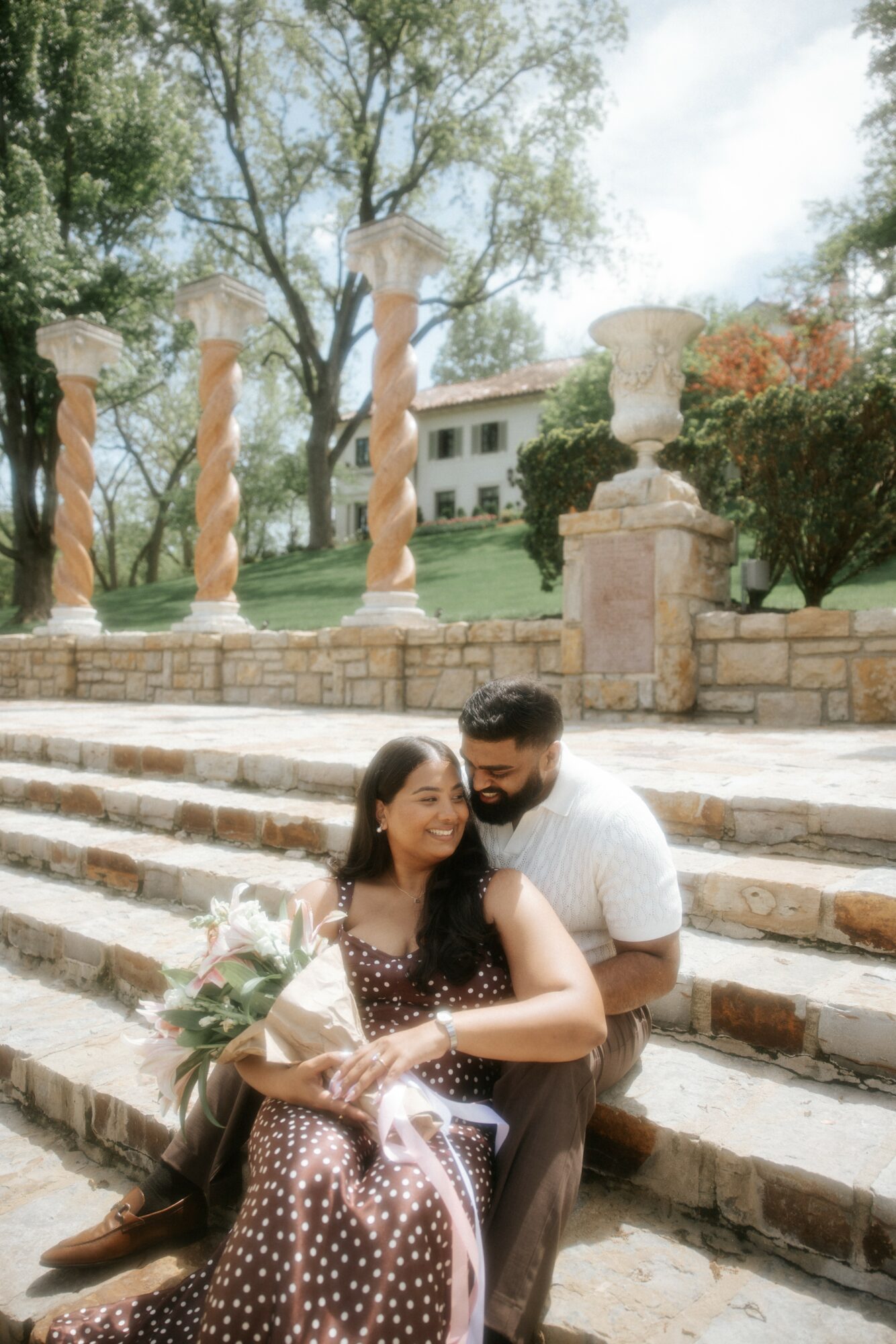 A couple sitting on stone steps outdoors, smiling, with trees and a building in the background.