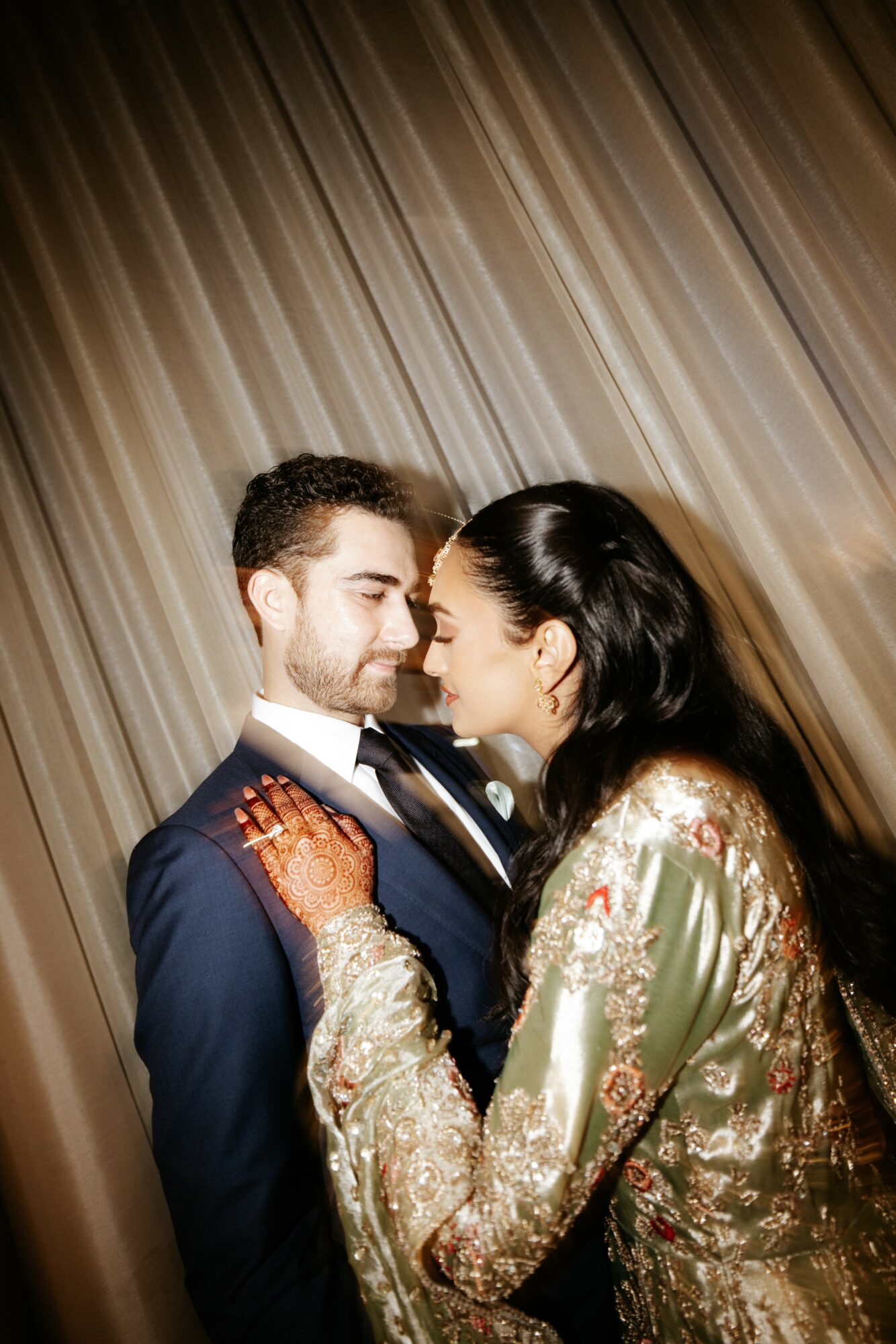 A couple faces close together, with the woman touching the man's chest, against a curtain background.