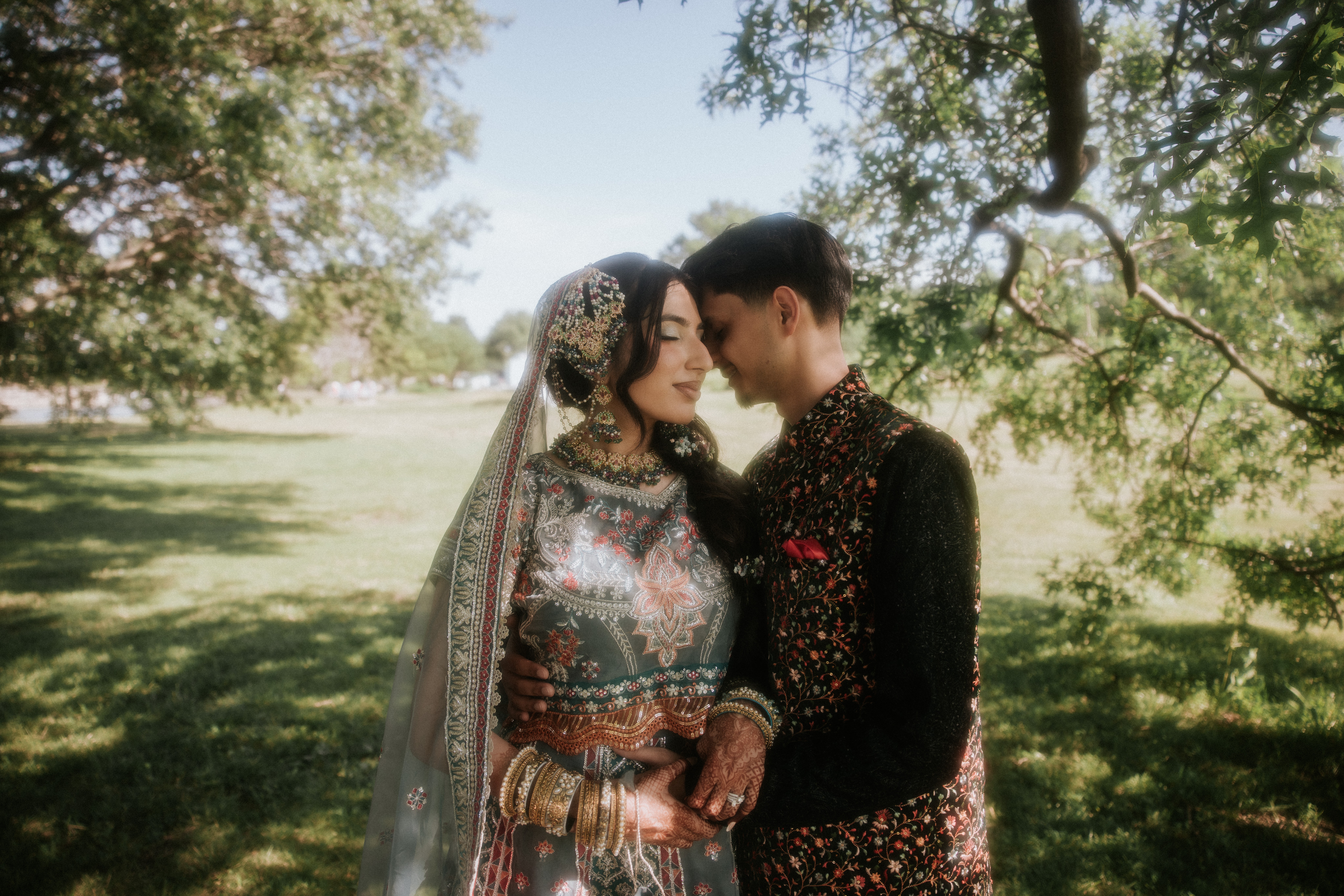 A couple in traditional attire standing close under a tree in a park, holding hands and looking at each other.