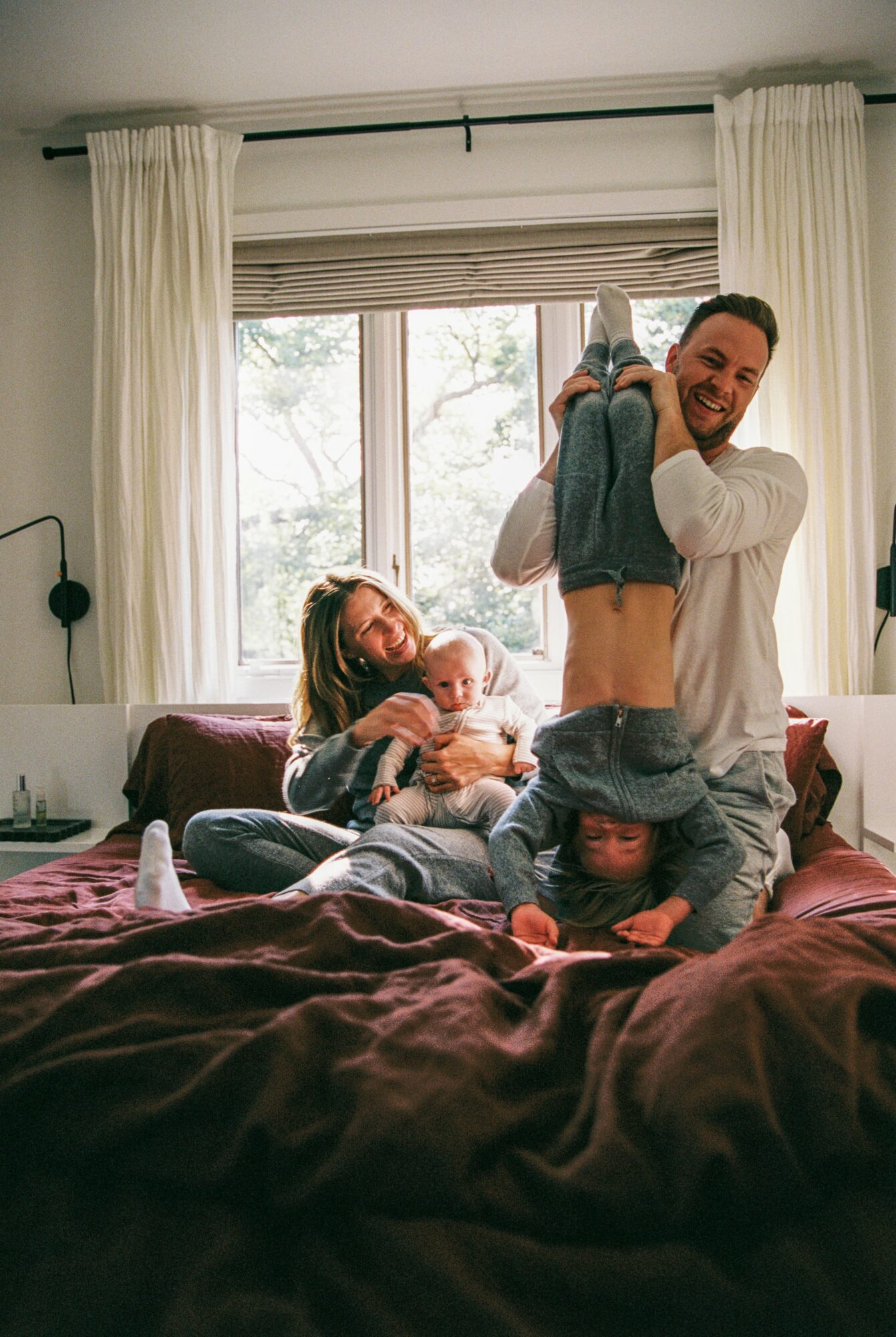 Family on bed, man lifting child upside down, woman holding baby, bright window behind, cozy room.