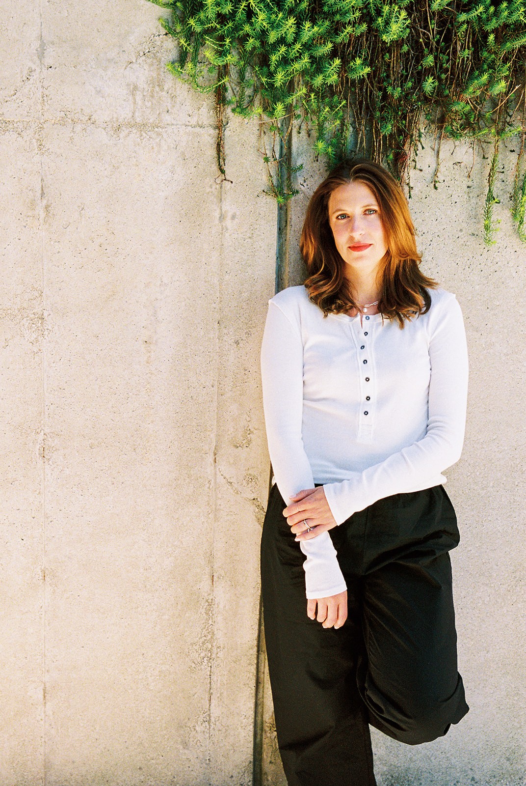 Woman with shoulder-length hair wearing a white long-sleeve top and black pants, standing against a concrete wall with greenery above.