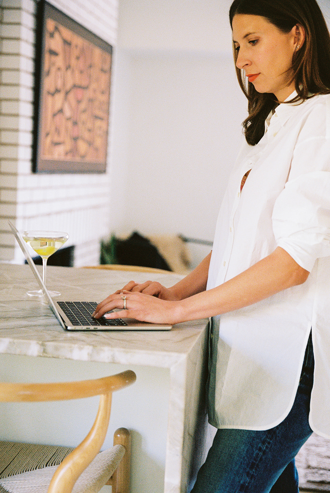 Woman in white shirt working on laptop at kitchen counter, with artwork on wall behind her.