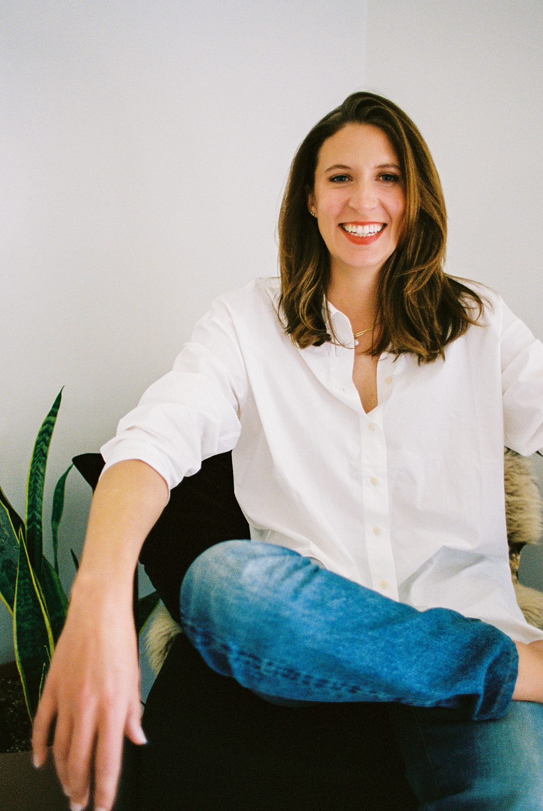 Smiling woman with shoulder-length hair wearing a white shirt, sitting on a chair with a plant nearby.