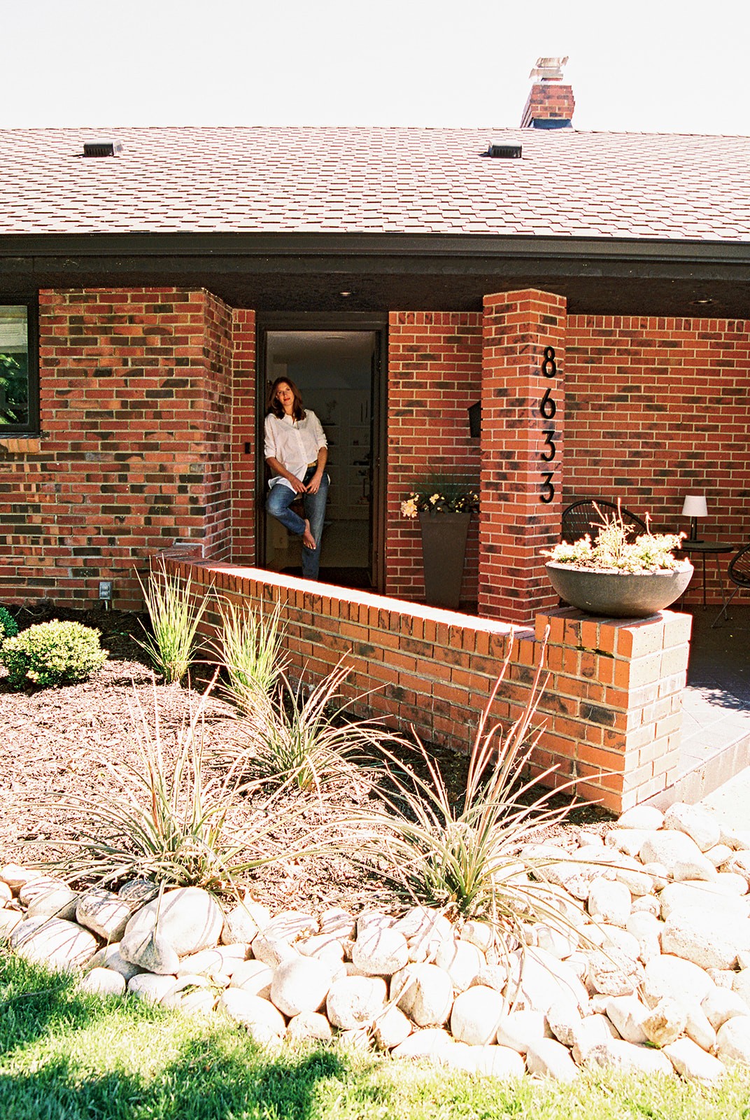 Person standing in doorway of brick house with garden and rocks in front, house number 8633 visible on brick pillar.