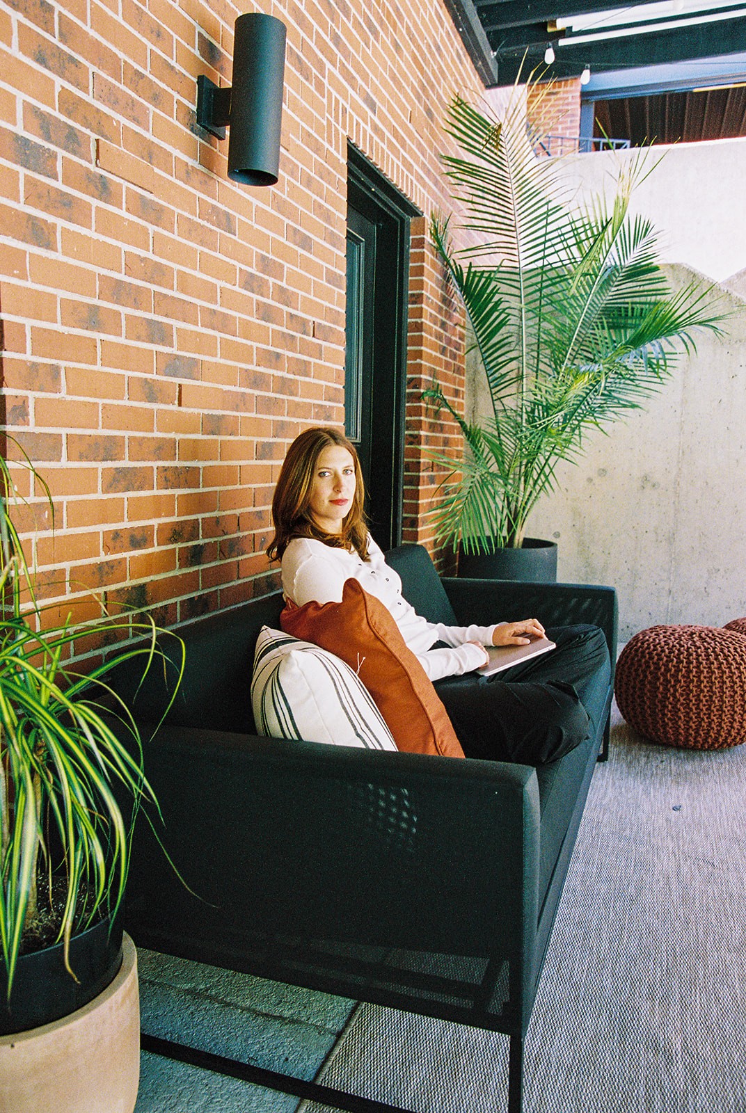 Woman sitting on a black sofa with pillows, next to large potted plants and a brick wall, outdoors or in a patio area.