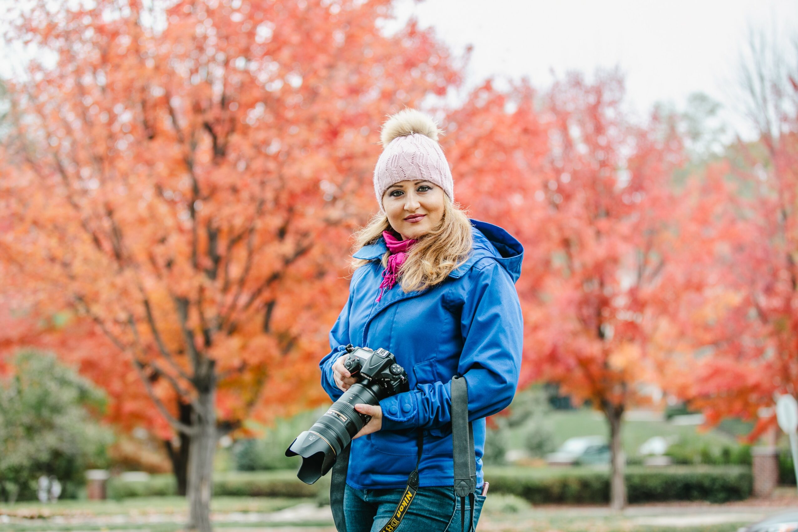 Woman in blue jacket holding camera outdoors with colorful autumn trees in background.