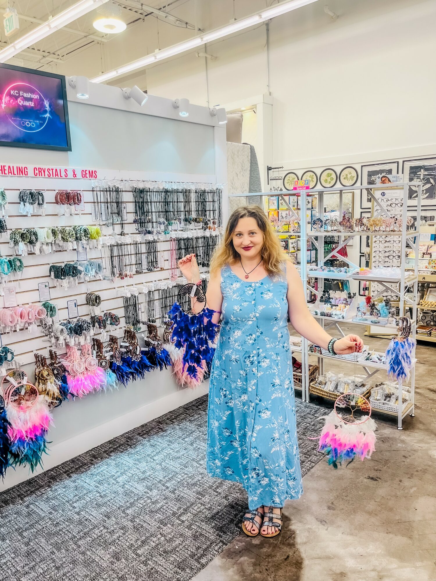 Woman in a blue dress shopping for accessories in a store, holding colorful feathered items, with shelves of jewelry and accessories behind her.