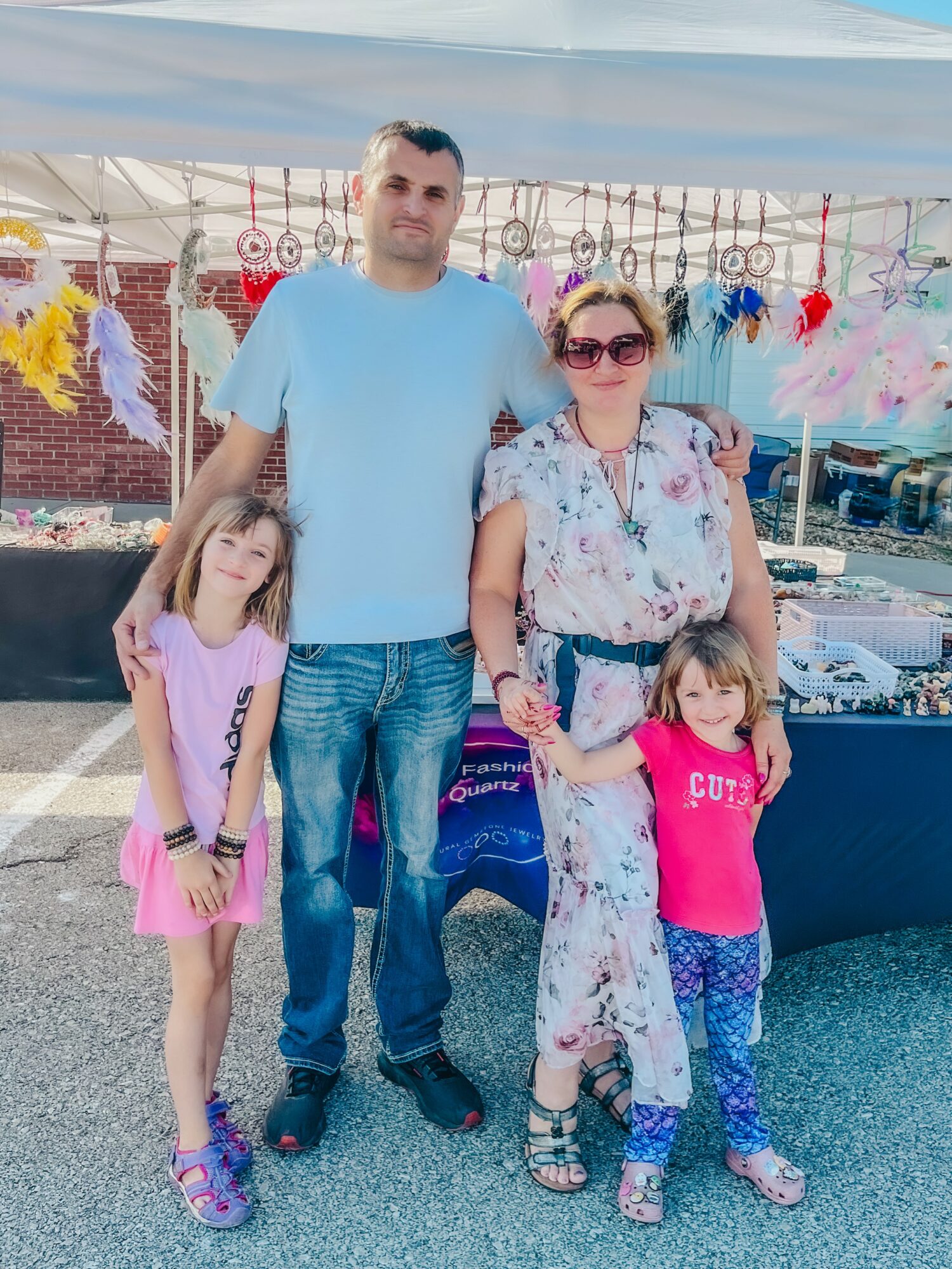 Family of four standing outdoors under a canopy, with colorful decorations in the background, smiling at the camera.