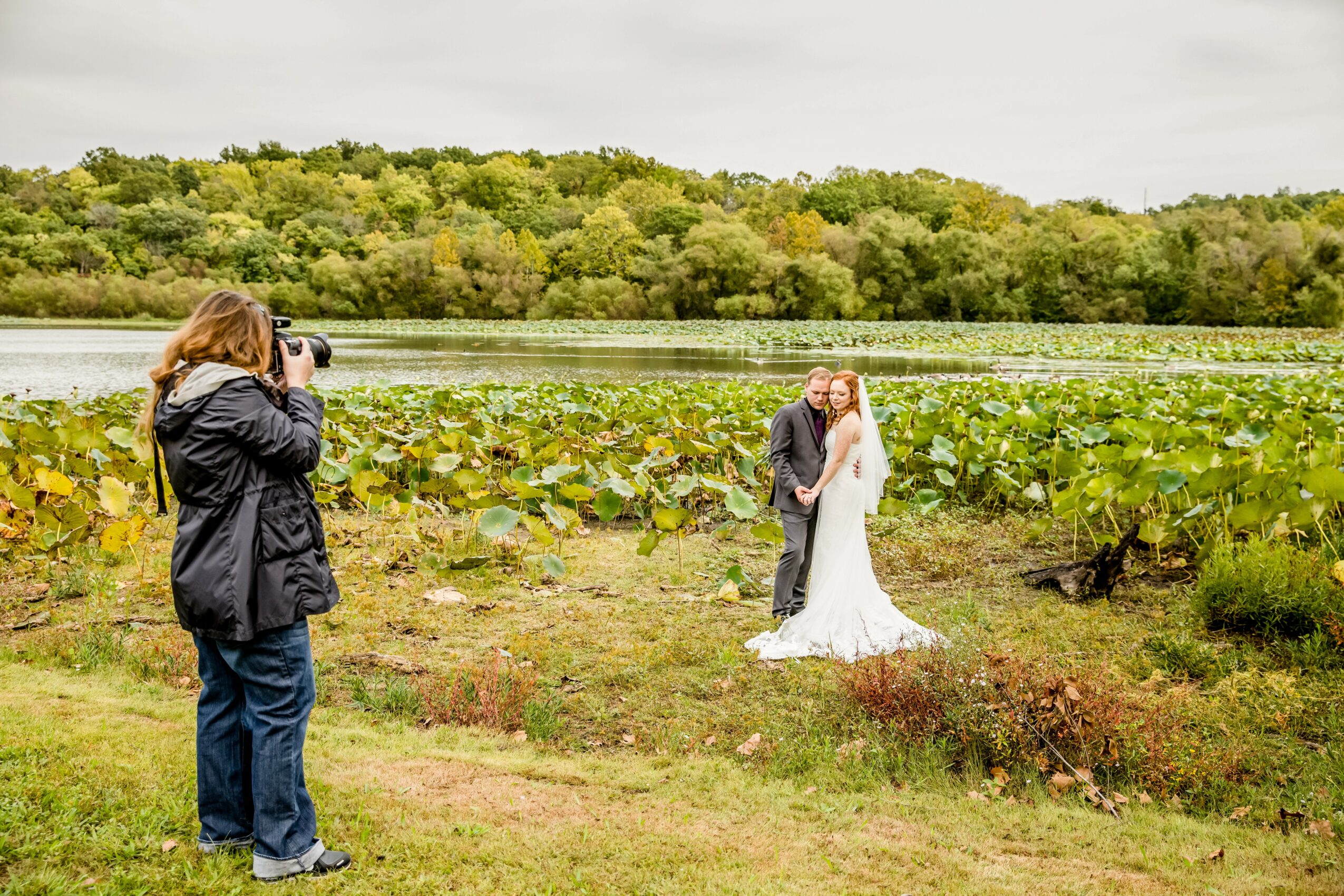 Two people taking a photo of a couple in wedding attire near a lake with green trees in the background.