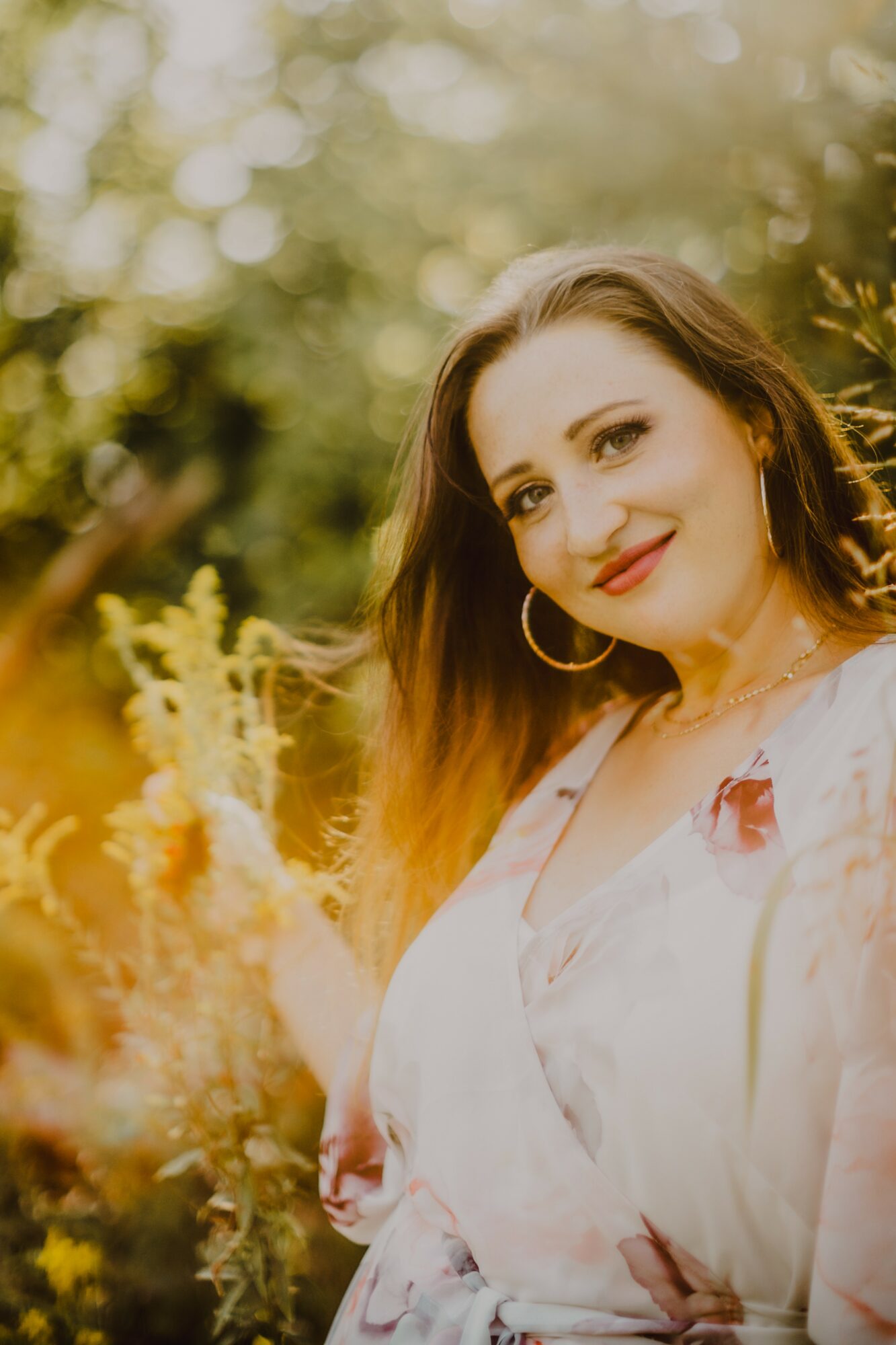 Smiling woman with long hair and hoop earrings outdoors, surrounded by yellow flowers and greenery.