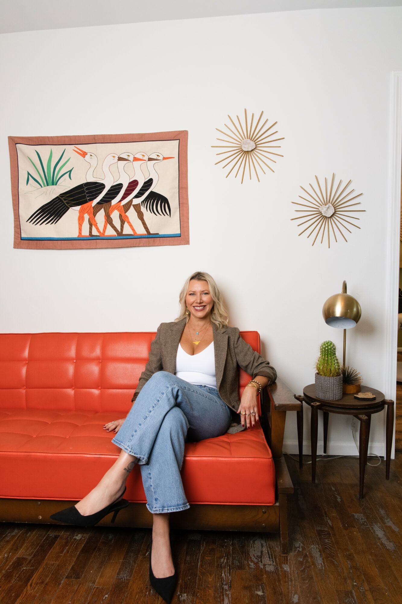 Woman sitting on a red sofa in a room with wall art and decorative wall hangings, smiling, wearing a blazer and jeans.