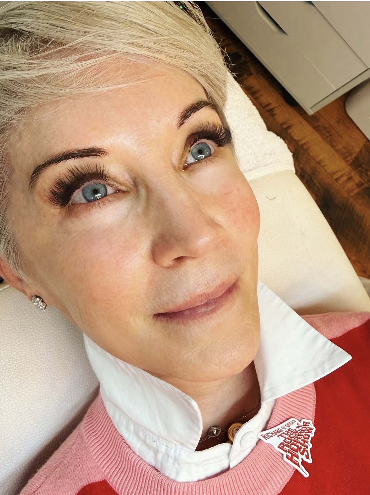 Close-up of a woman with blonde hair and blue eyes, lying on a white surface, wearing a white collared shirt and red sweater.