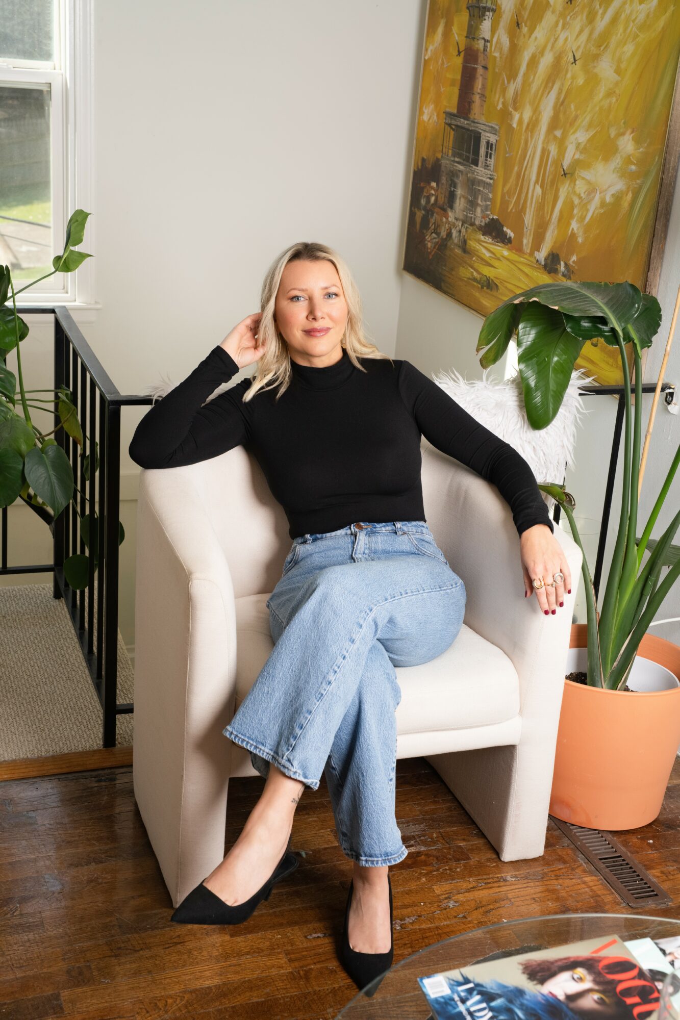 Woman with blonde hair in black top and jeans sitting on a white armchair, in a room with plants and artwork.