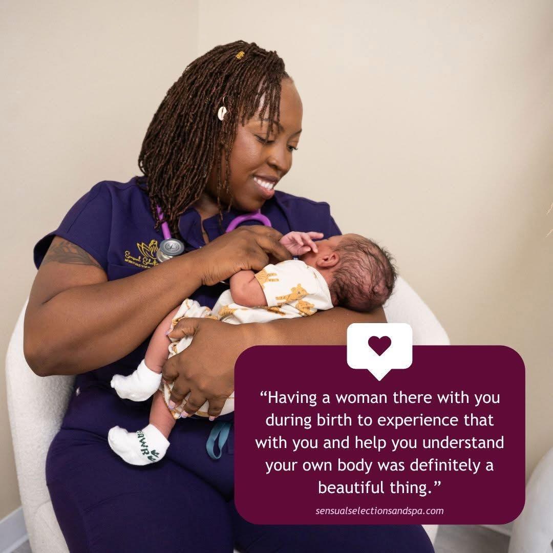 A woman in a purple uniform holds a newborn baby, smiling at the infant against a plain background.