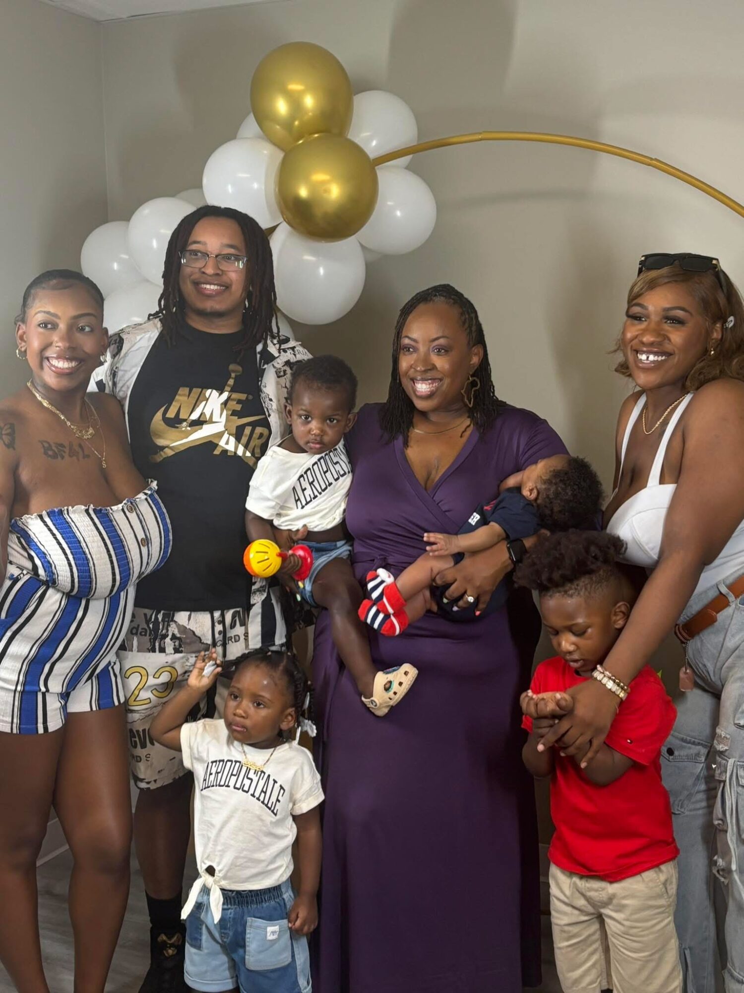 Group of women and children smiling, standing in front of white and gold balloons, celebrating indoors.
