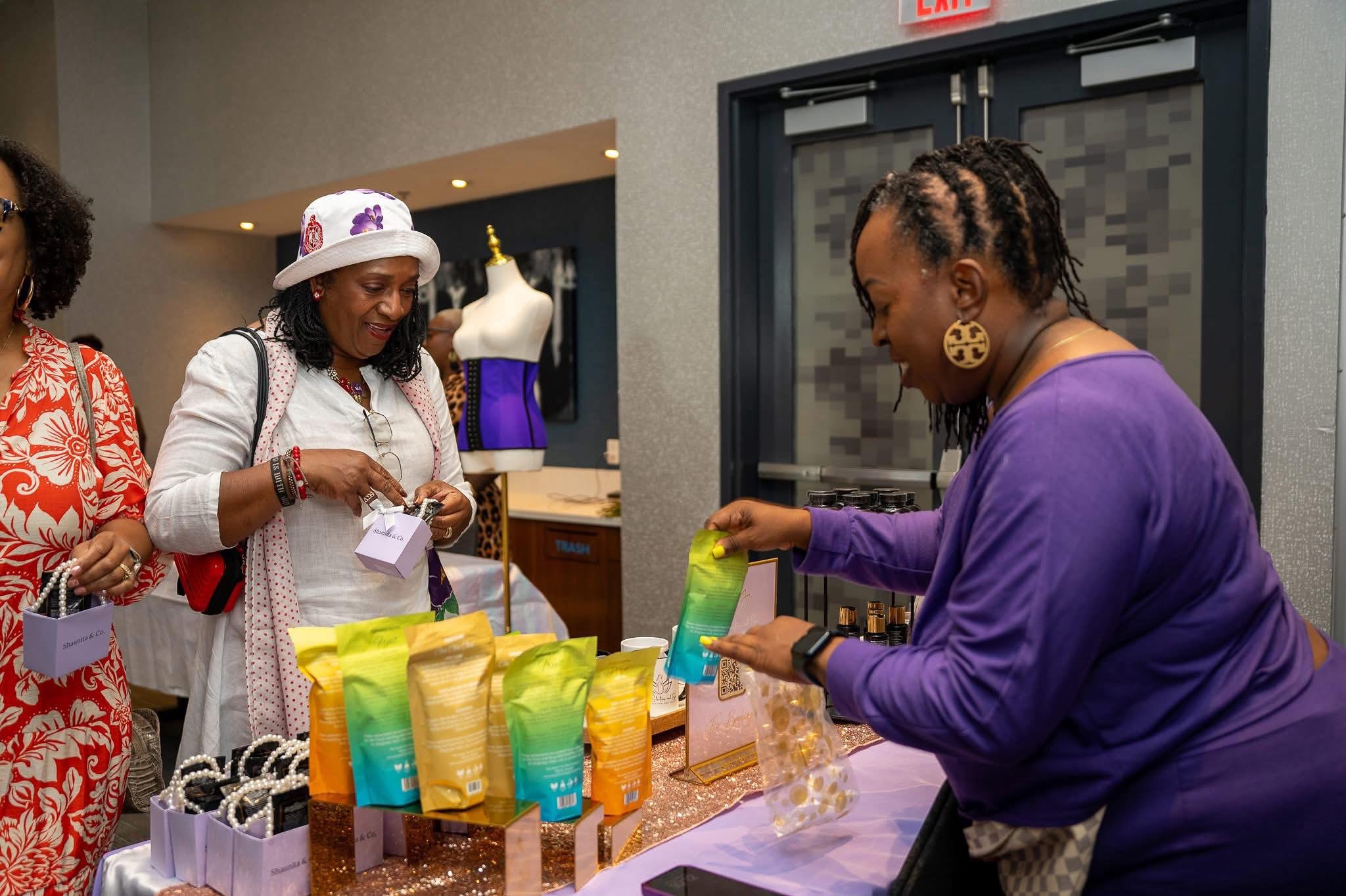 Woman in purple shirt arranging colorful gift bags on table, others in background at indoor event, mannequin in store window.