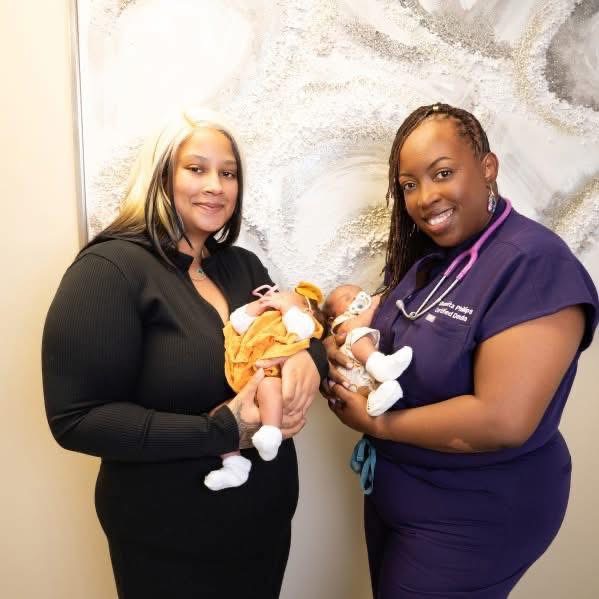 Two women holding newborn babies, standing indoors against a textured wall, smiling at the camera.