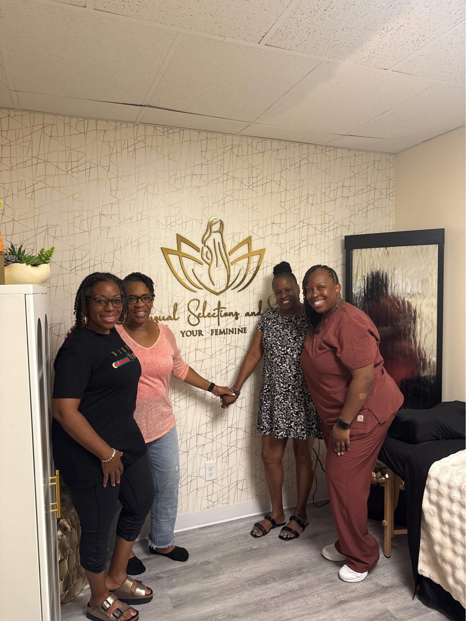 Four women standing together in a room, holding hands, smiling, with a wall decoration and framed picture behind them.