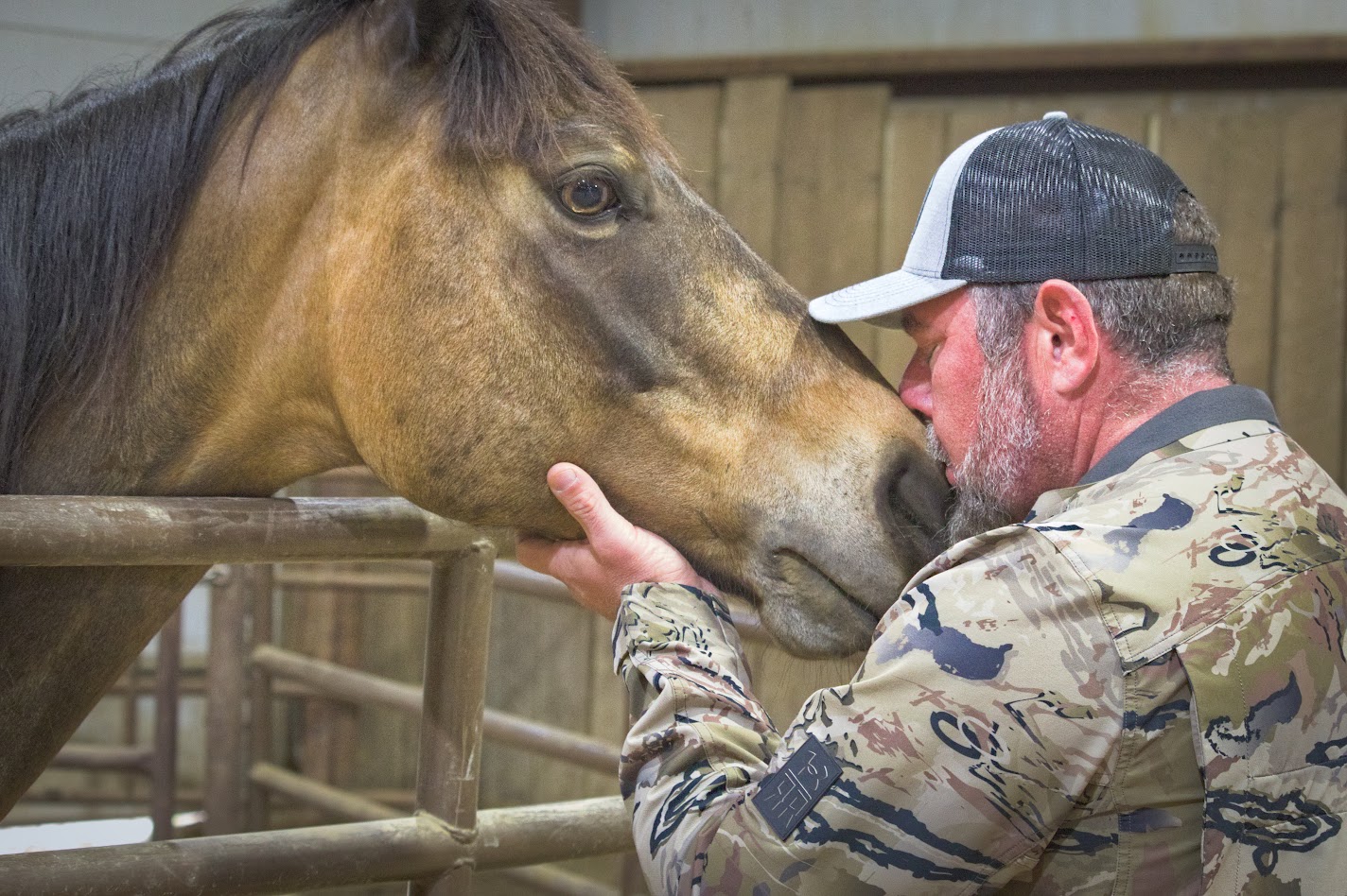 Man in camouflage jacket and cap gently touching a horse's nose inside a wooden enclosure.