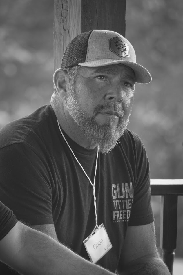 Man with a beard and cap sitting outdoors, wearing a T-shirt and a lanyard, looking to the side.