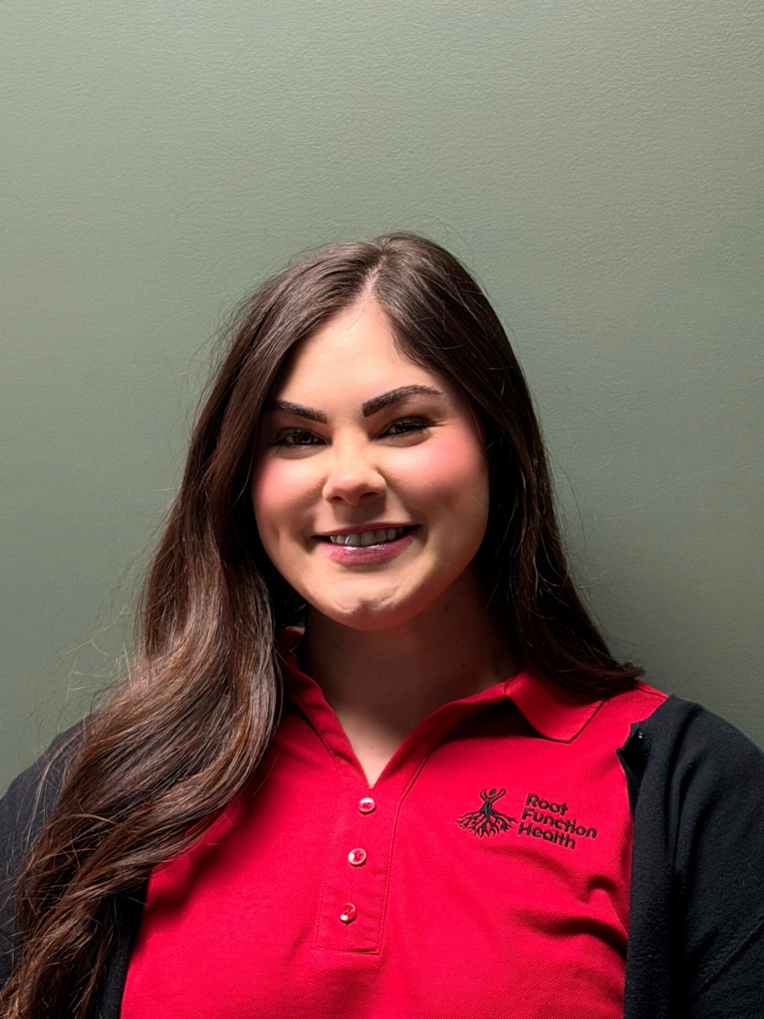 Young woman with long brown hair smiling, wearing a red polo shirt with a logo, standing against a plain green background.