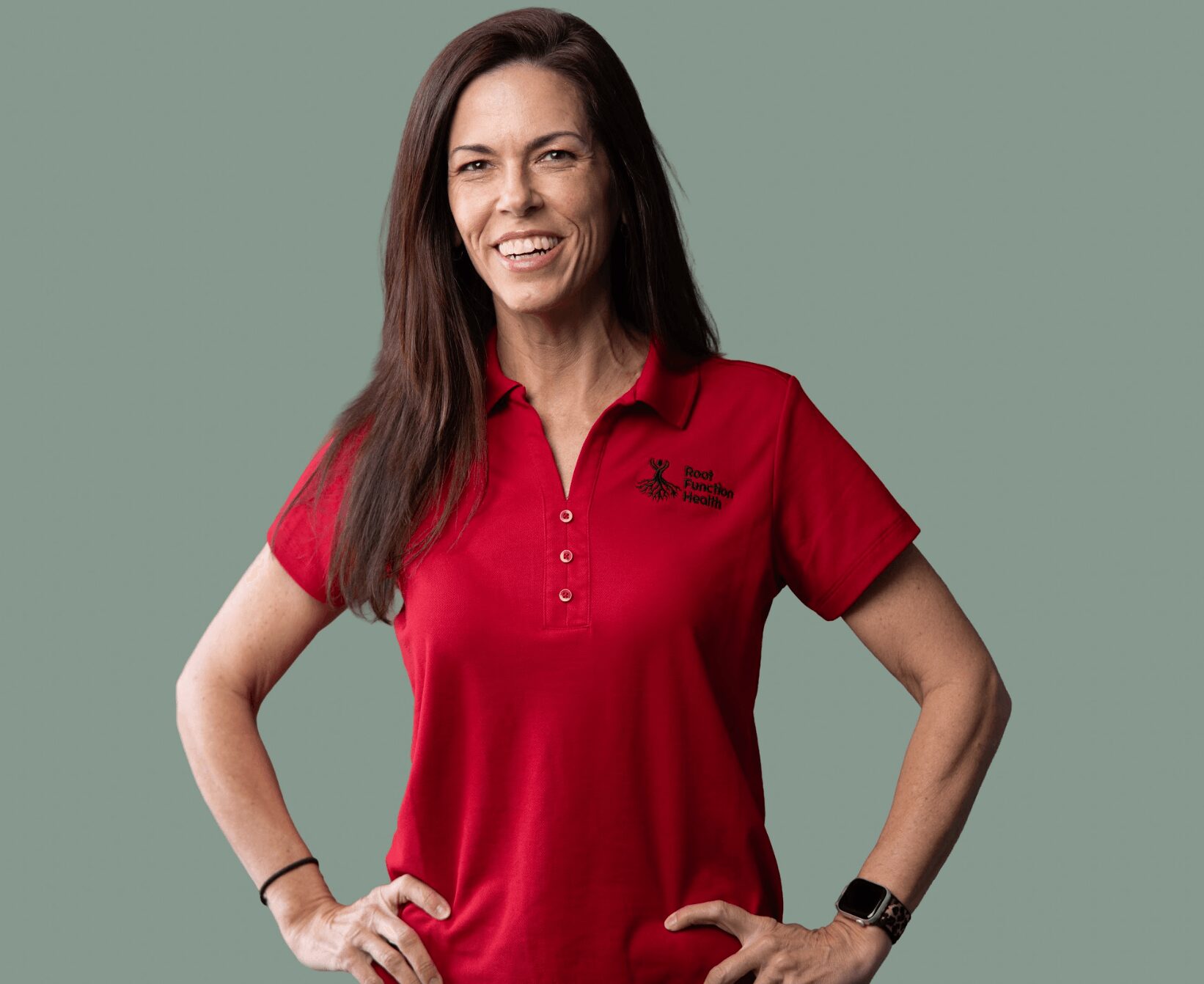 Woman with long dark hair wearing a red polo shirt, smiling, hands on hips, against a muted green background.