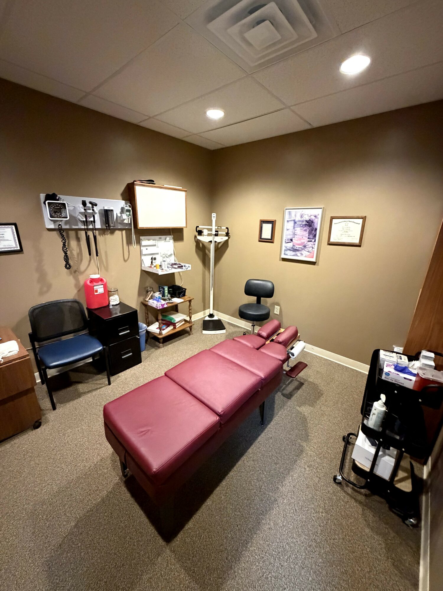 Medical examination room with a red examination table, chairs, and medical equipment on the walls.