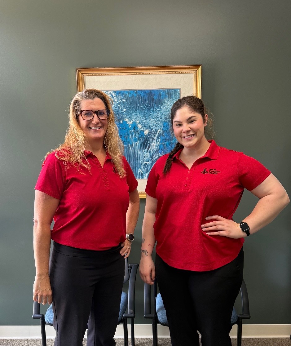 Two women in red shirts standing indoors in front of a framed artwork, smiling, with chairs behind them.