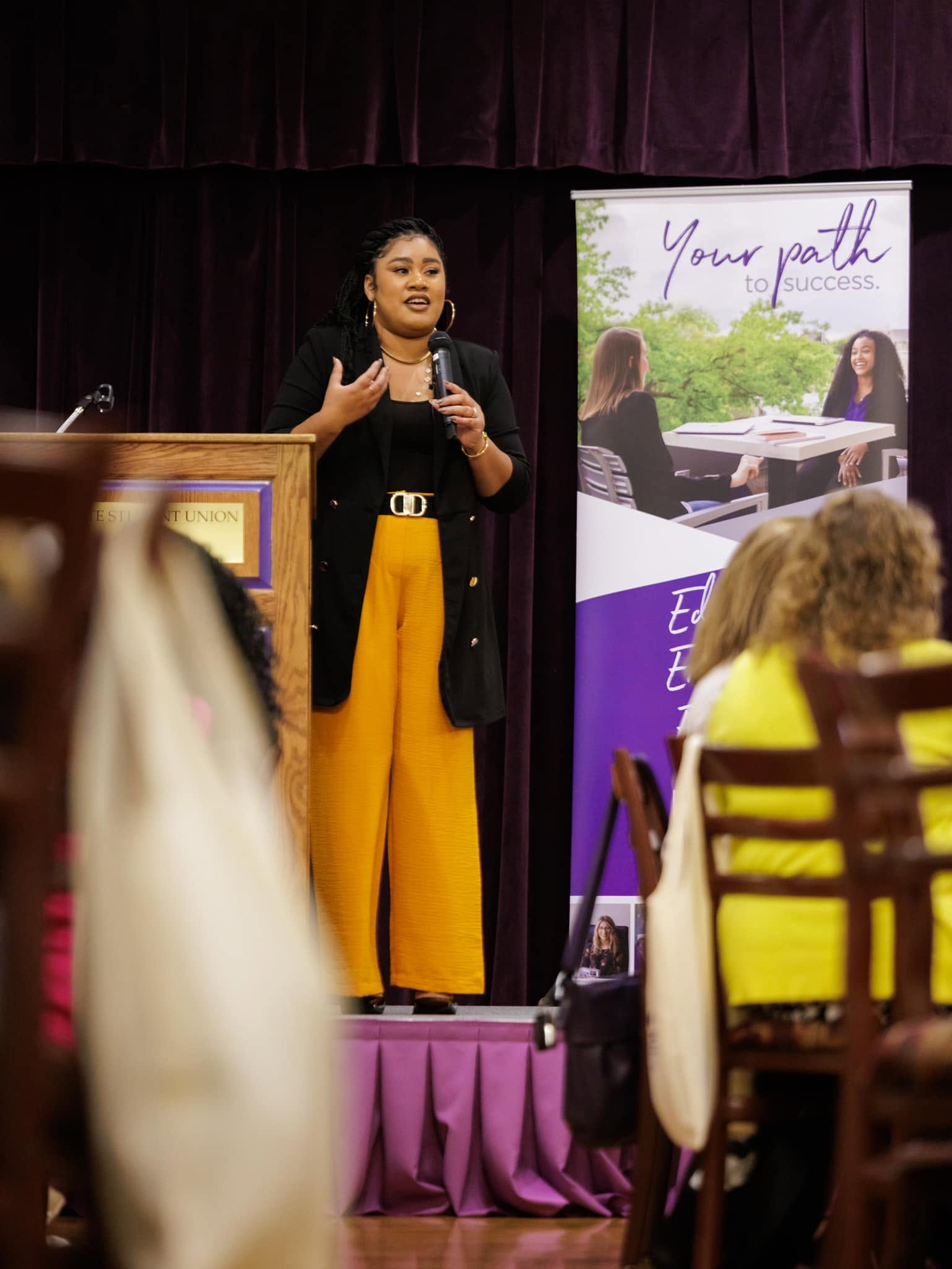 Woman speaking on stage with a microphone, audience seated, purple curtain backdrop, and a banner with text and images.