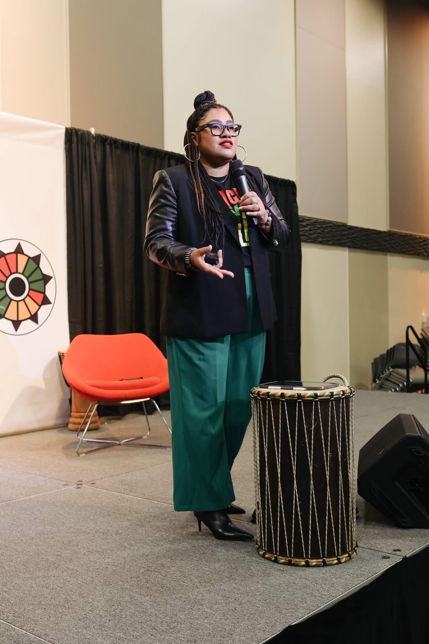 Woman speaking into microphone on stage, wearing glasses, black jacket, green pants, with a drum nearby.