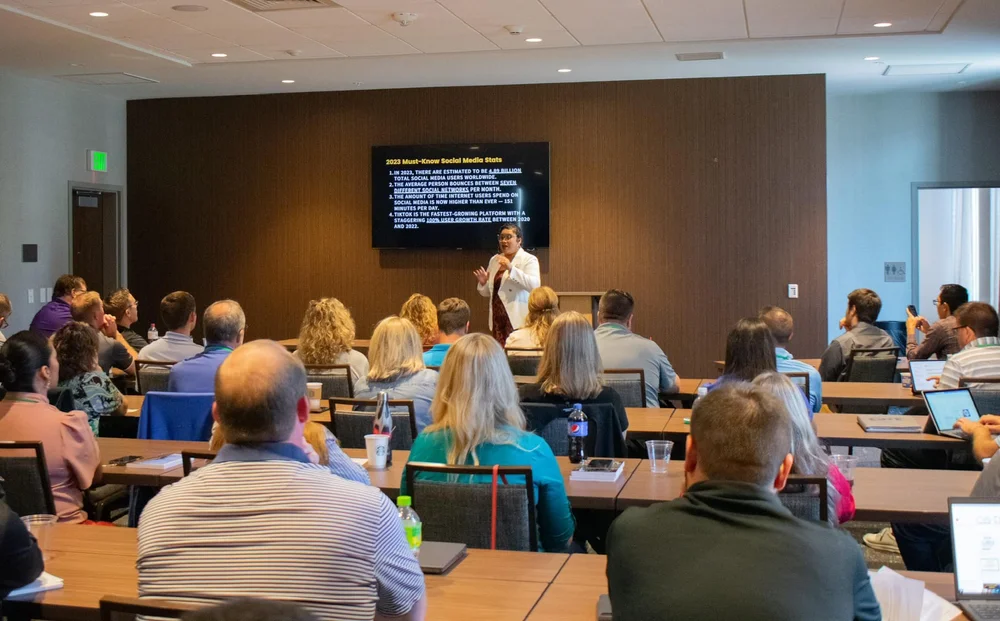 A presenter stands in front of a screen in a conference room filled with seated attendees, some using laptops.